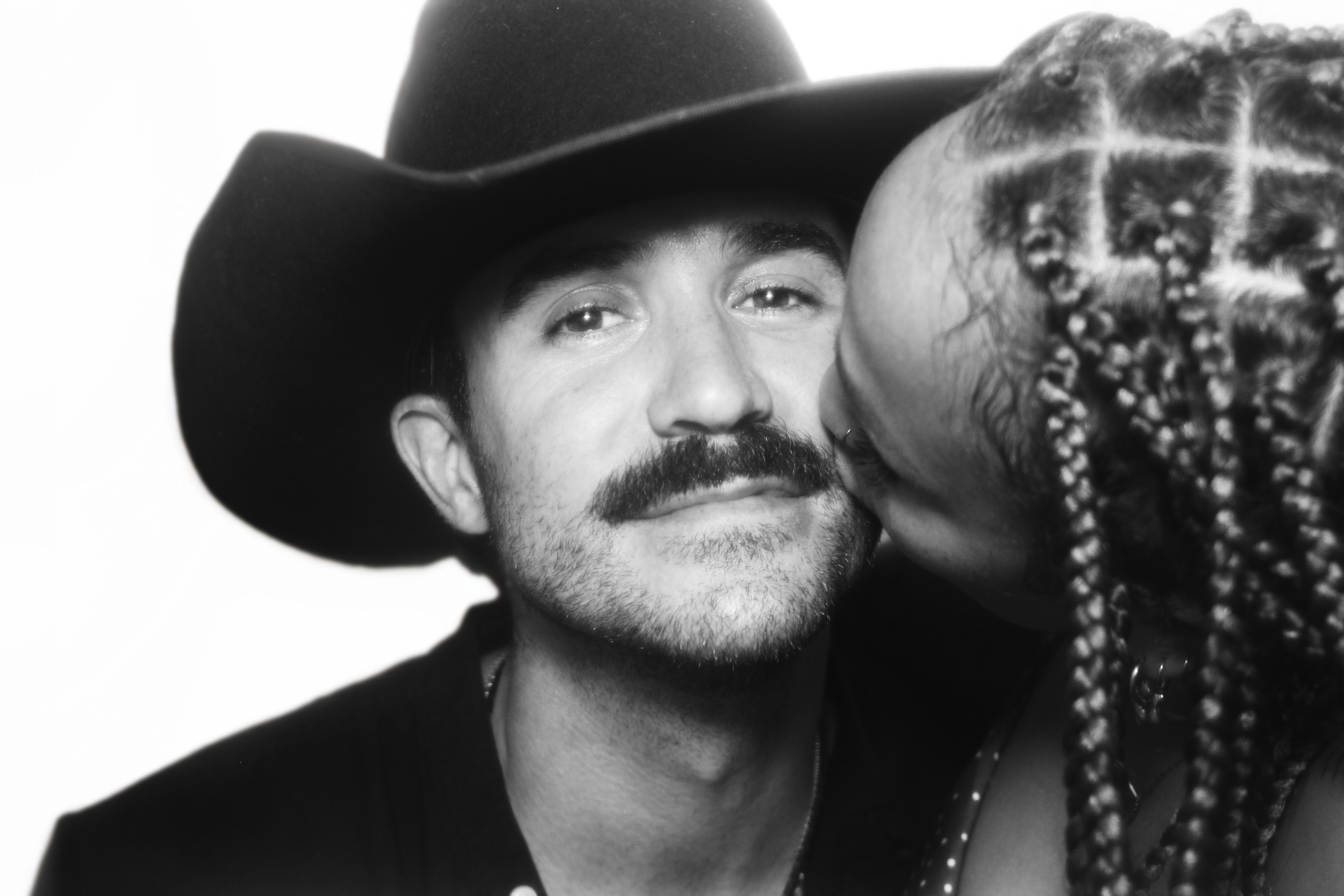 A man with a mustache and beard wearing a cowboy hat is being kissed on the cheek by a woman with braided hair in a photo booth.