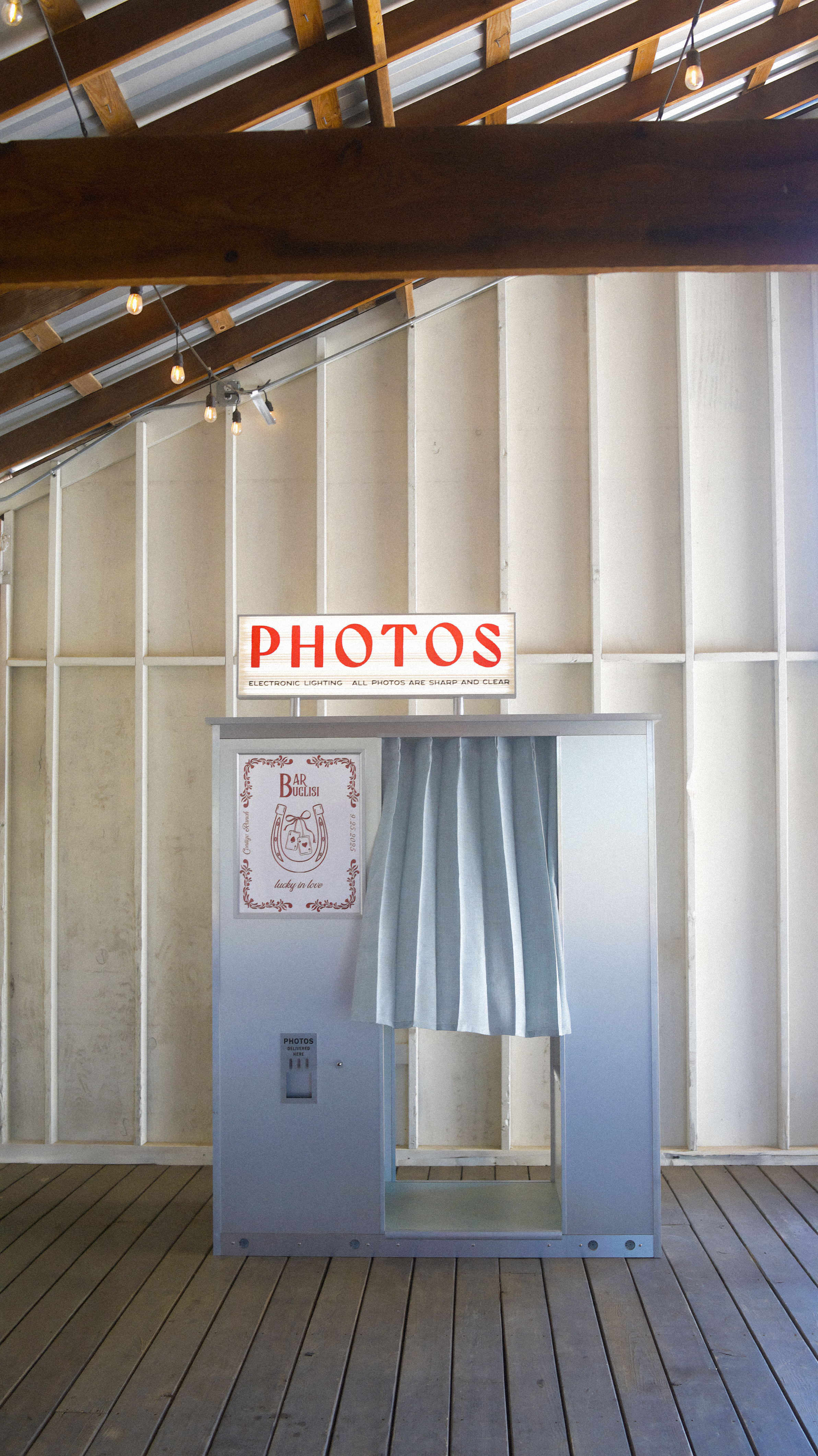 Photo booth with a gray structure, blue curtain, and a sign that reads 'PHOTOS'. The background, a Texas barn wedding venue, features a wall with wooden framing and a sloped ceiling with exposed beams and string lights.