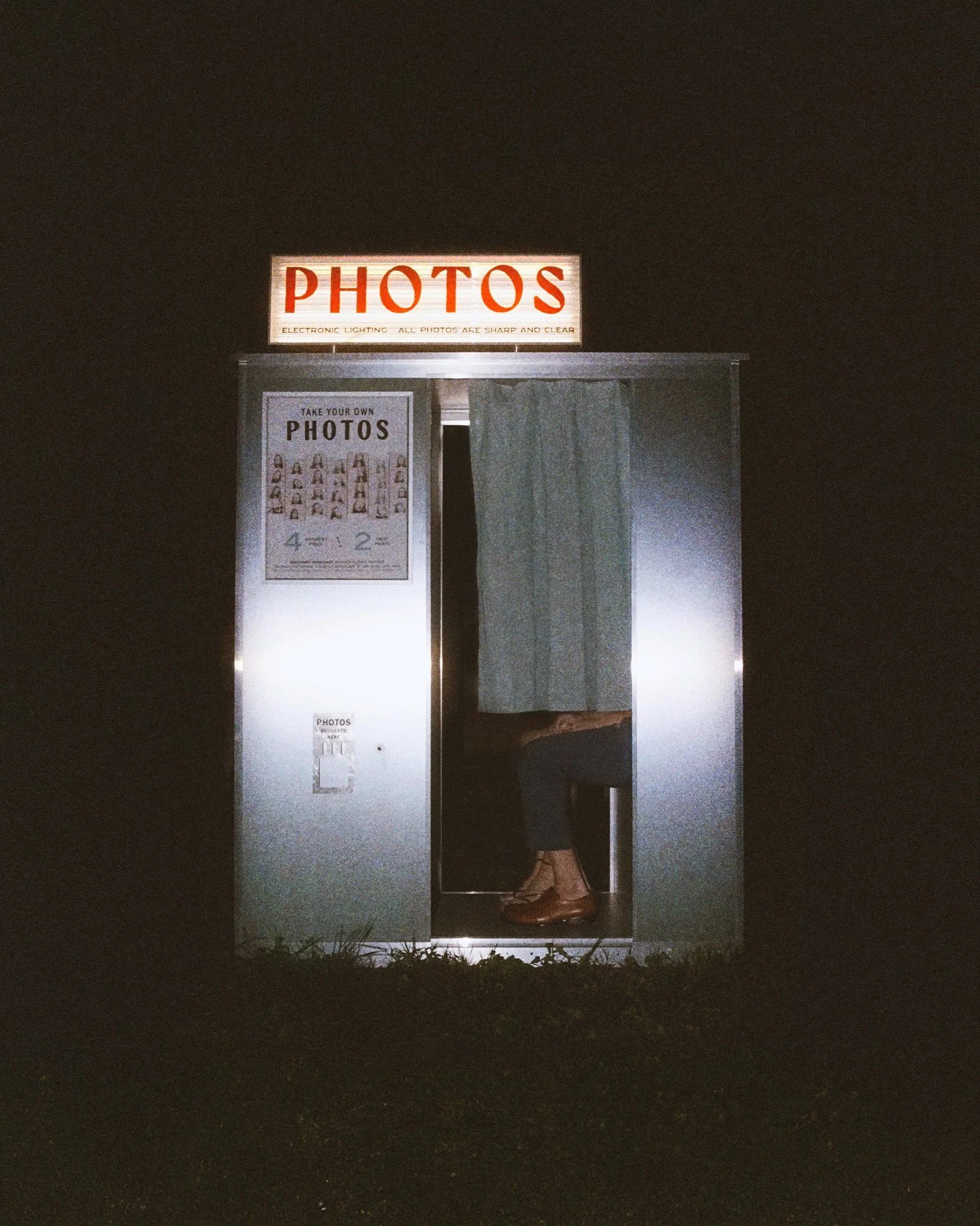 Vintage-style photo booth with a person sitting inside, partially visible, with the sign 'PHOTOS' on top, illuminated at night.