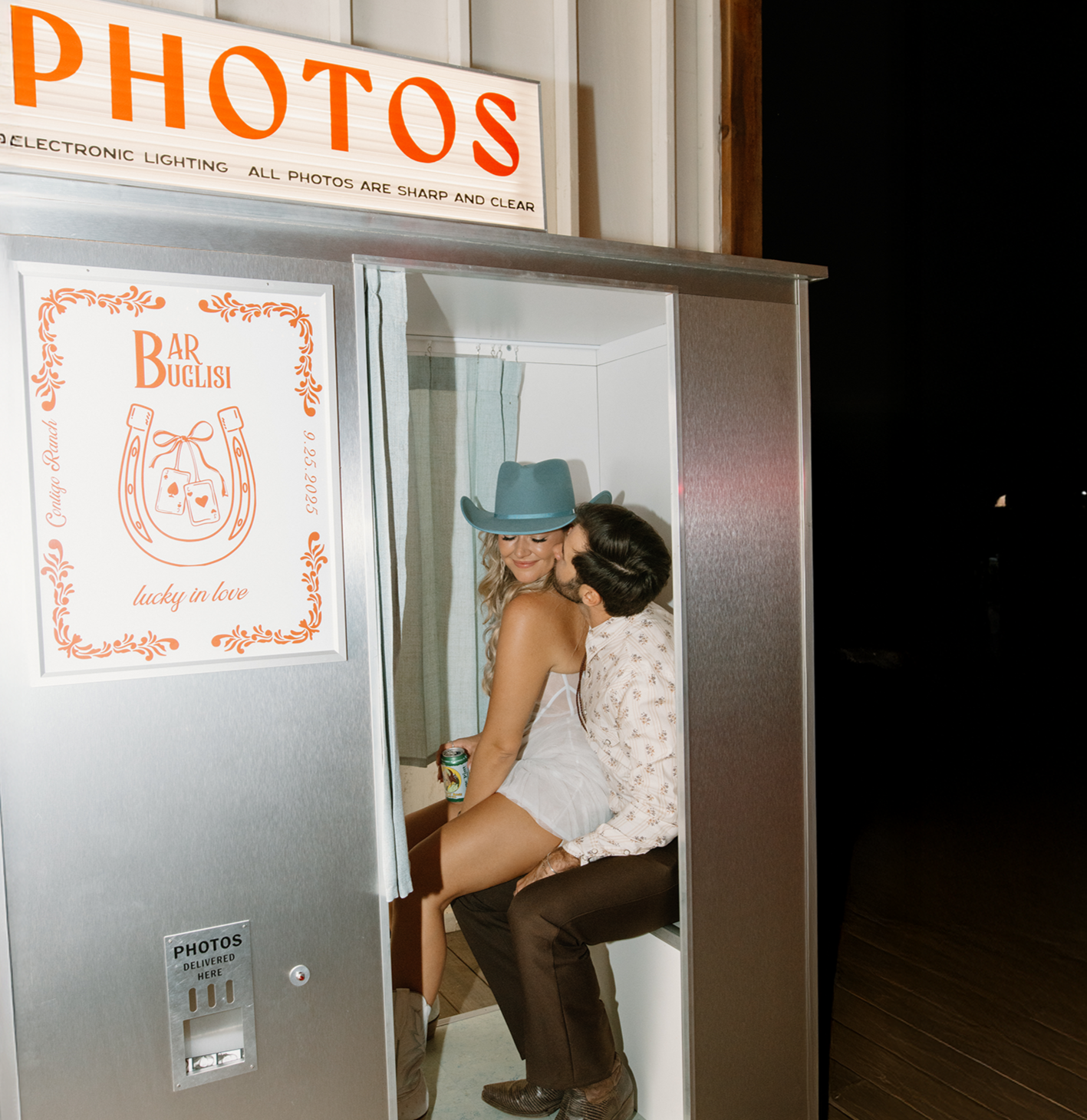 A couple sharing a tender moment inside a photo booth, with the woman smiling and wearing a large blue cowboy hat, and the man kissing her cheek. The photo booth is labeled "PHOTOS" and has a sign with decorative orange borders and text advertising a photo service.