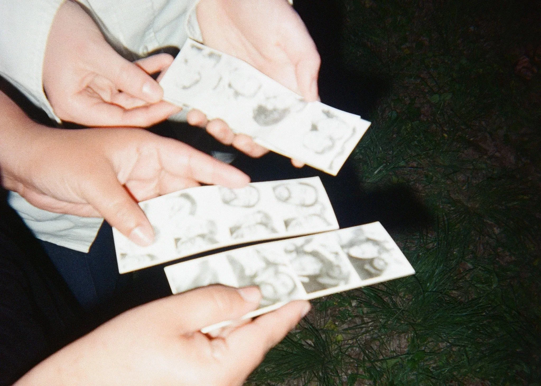 Close-up of two pairs of hands holding film photo strips with several black and white photographic images on them, with a dark, grassy background.
