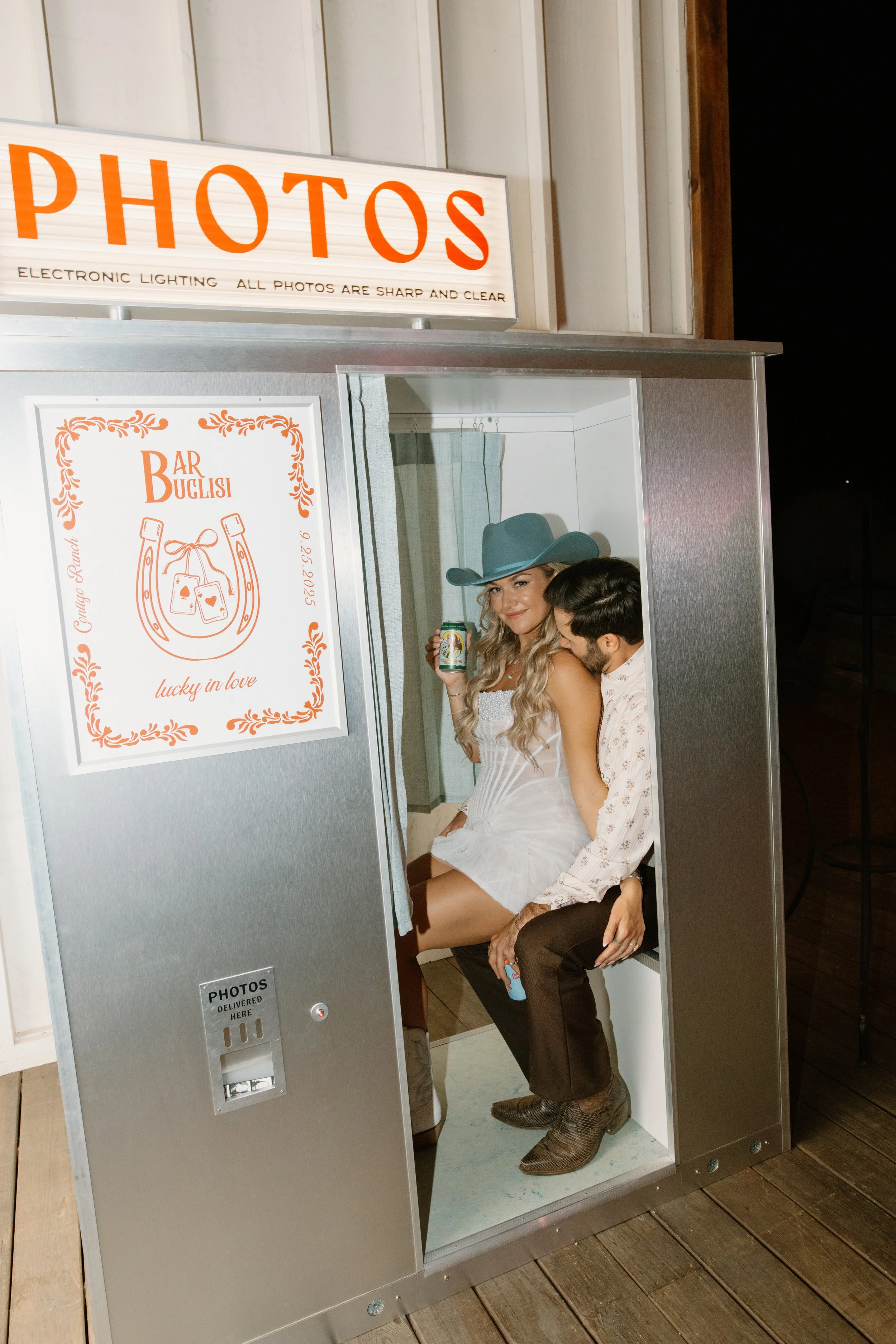 Two people sitting inside a photo booth at a wedding or event, one woman wearing a cowboy hat and holding a drink, the other person hugging her from behind, both smiling.