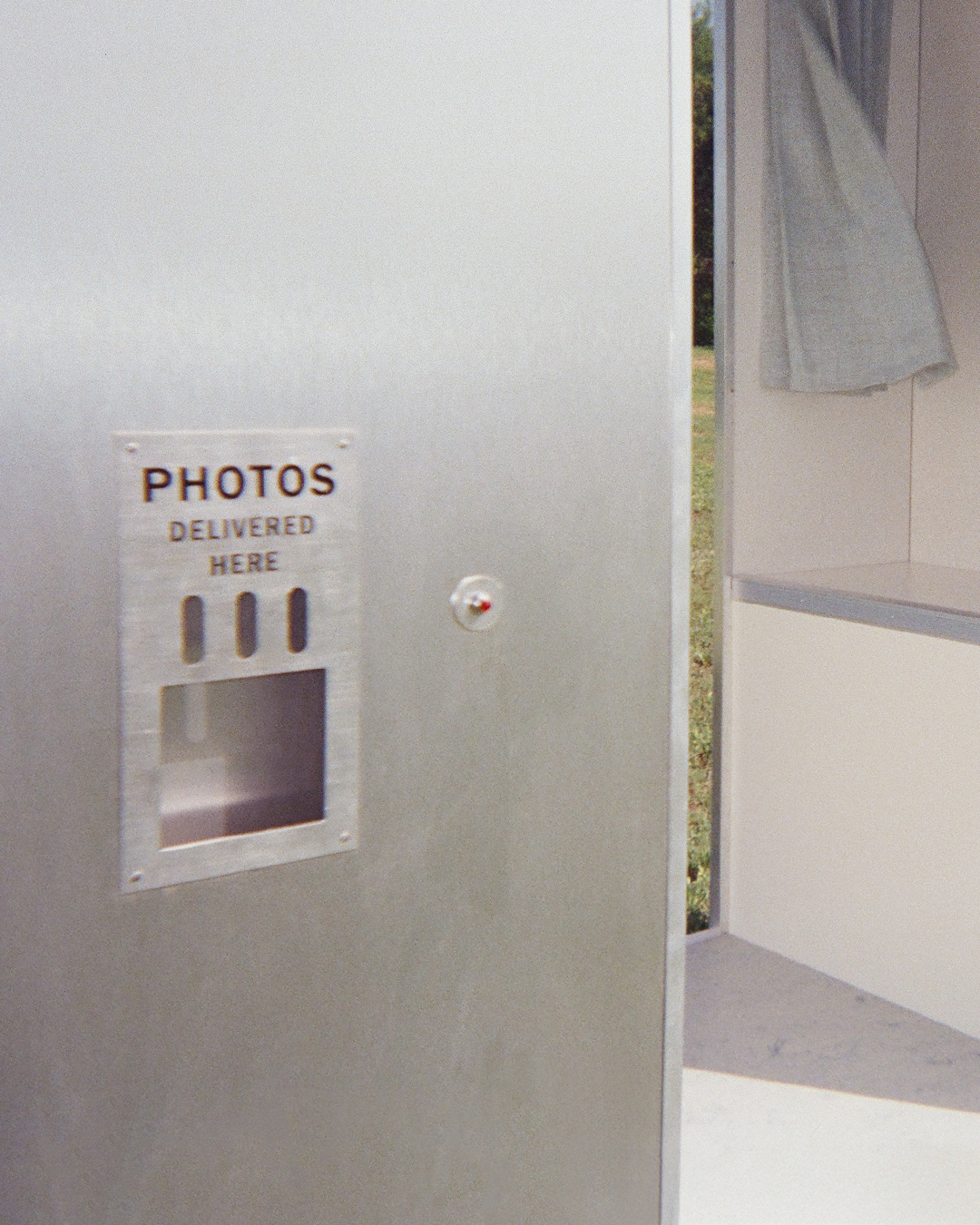 Photo of a photo booth drop box labeled 'Photos Delivered Here', located outside a vintage style photo booth with a curtain visible.
