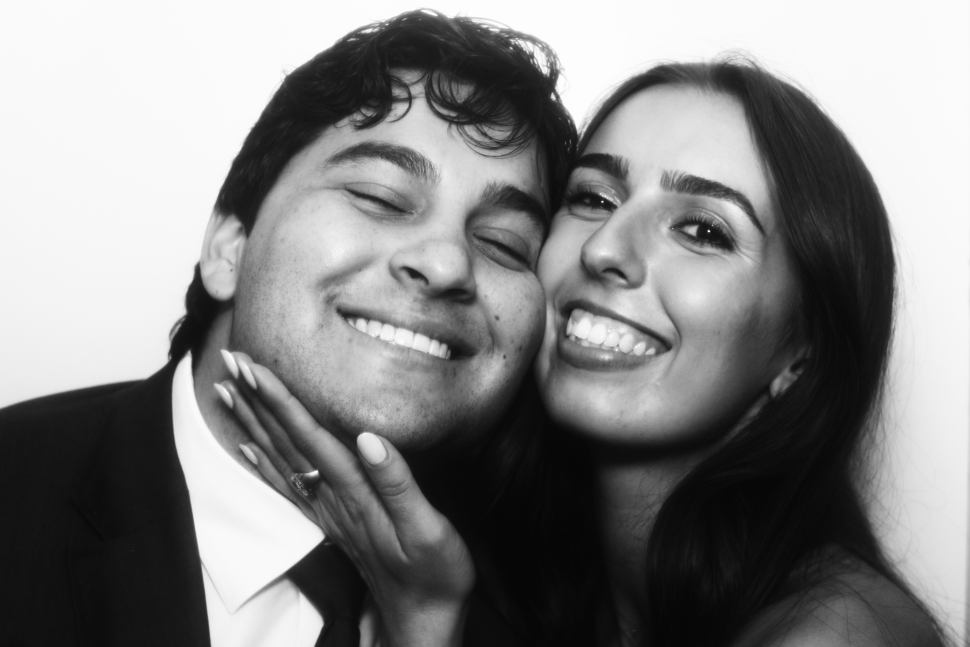 A black and white close-up photo of a smiling couple embracing inside of a vintage photo booth.