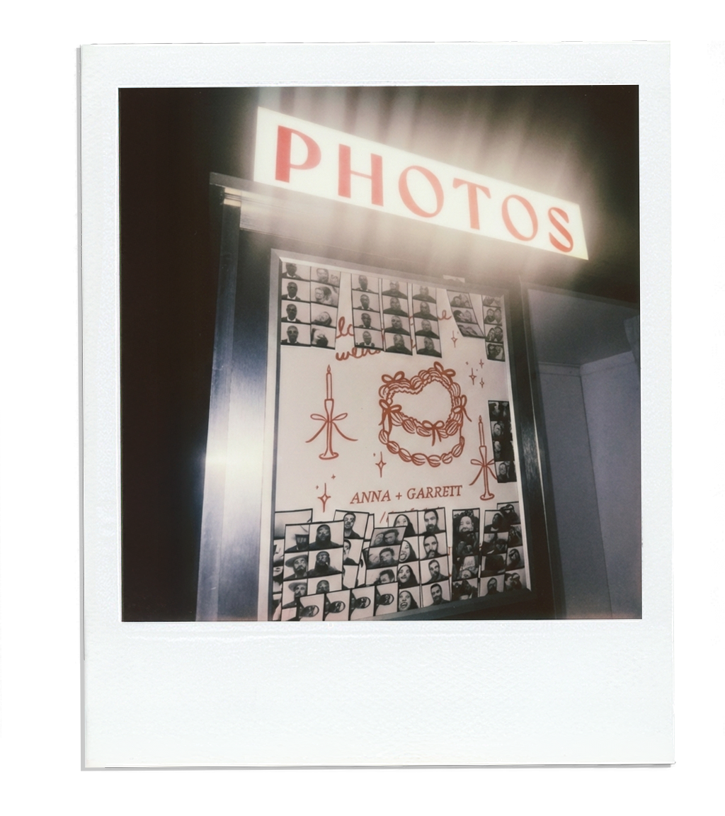 A photo booth display board with black-and-white photos of people, the names Anna and Garrett, candles, and a floral wreath, placed under a lit sign reading 'PHOTOS'.
