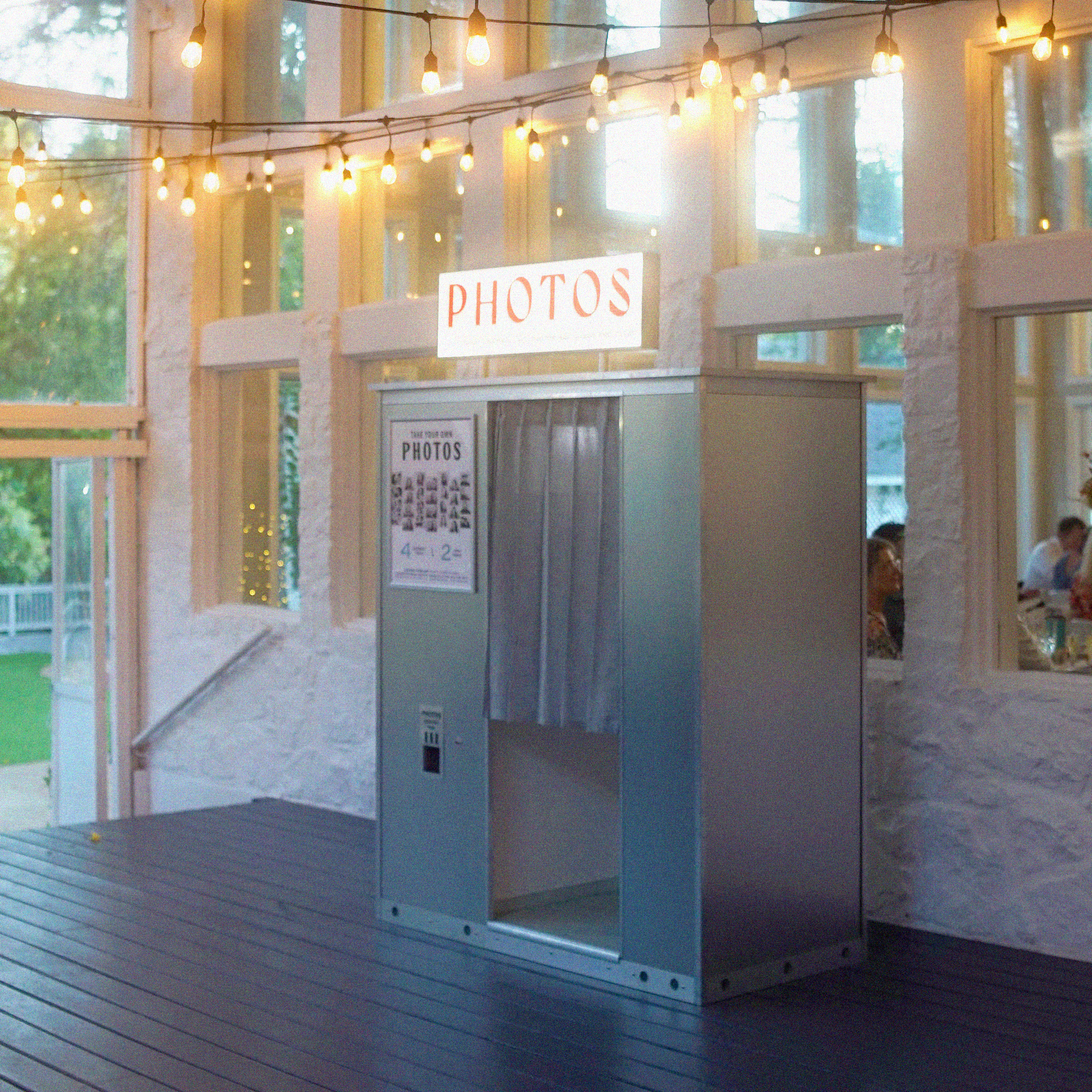 Photo booth with a  sign reading 'PHOTOS' inside a bright wedding venue with string lights and large windows.