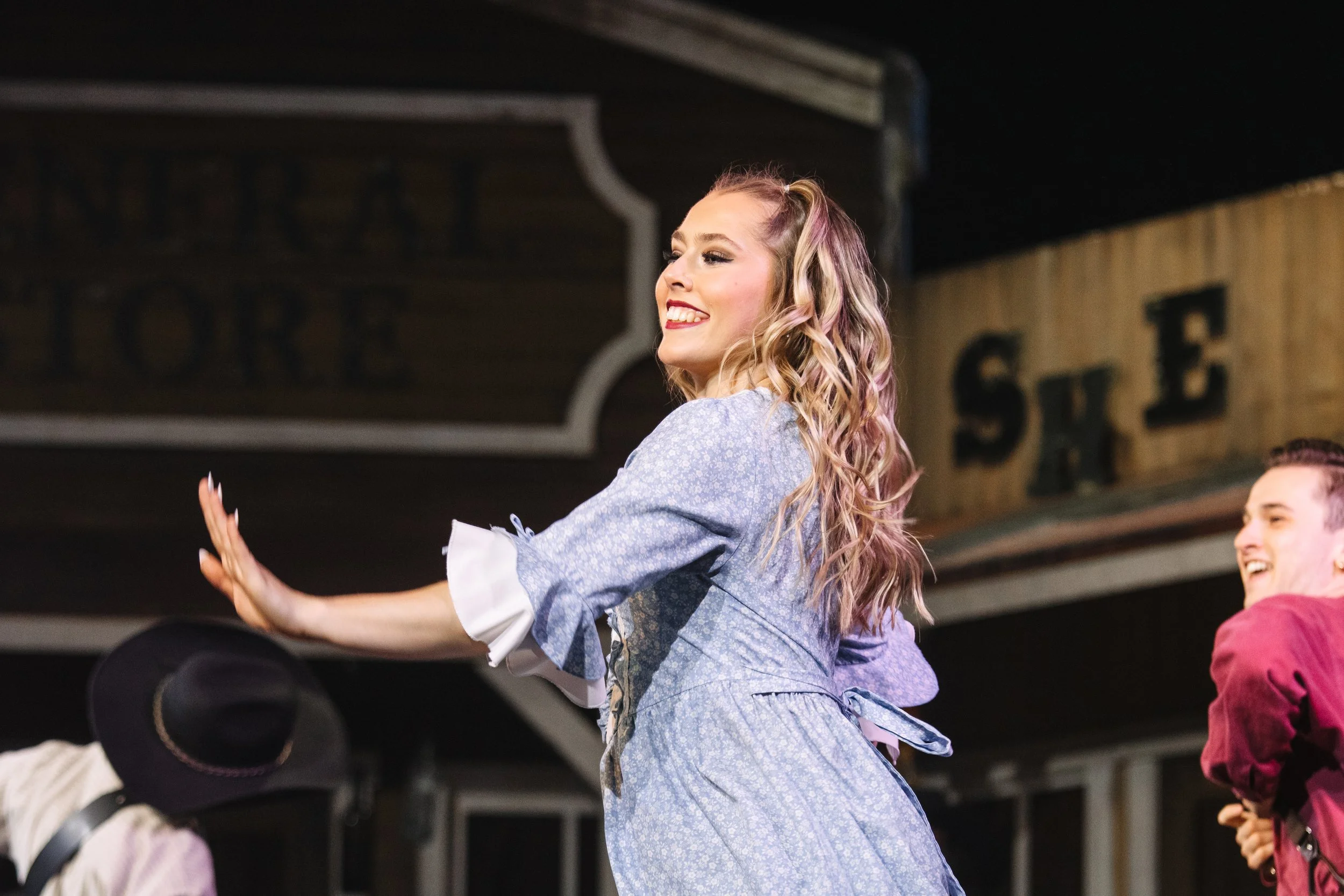 A young woman with long, wavy blonde hair, smiling and dancing, wearing a blue dress with ruffled sleeves, in a lively indoor setting with other people around.
