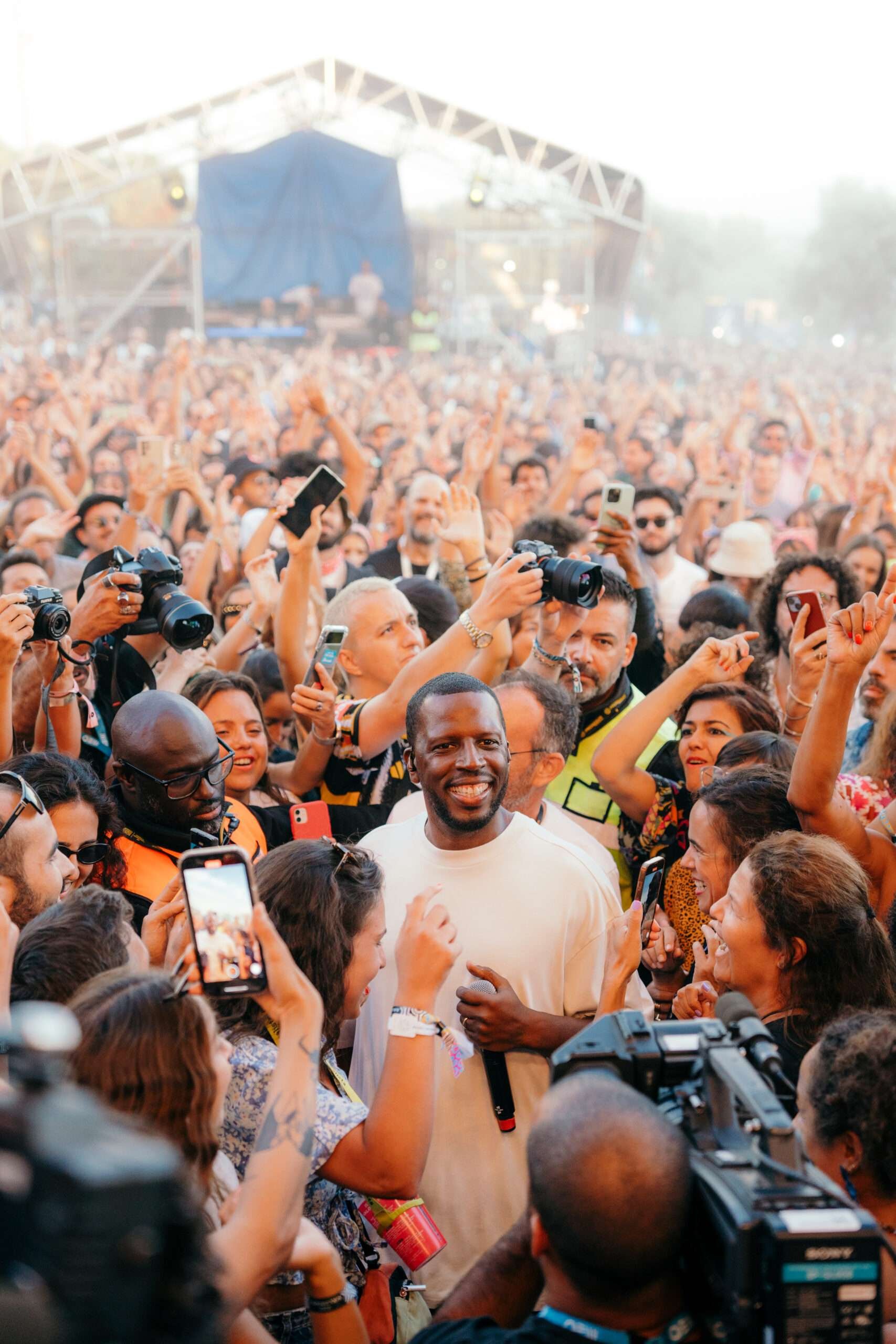 Pessoa sorridente cercada por uma multidão em um evento ao ar livre, com várias pessoas tirando fotos e filmando.