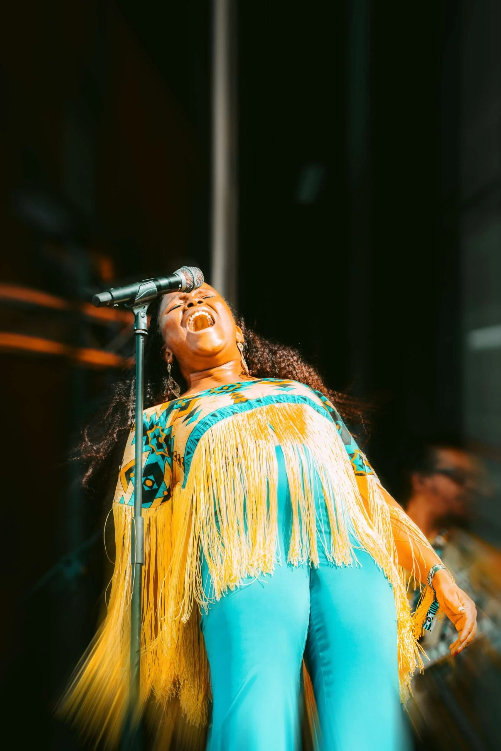 Mulher cantando com expressão de alegria, usando roupa colorida de cultura brasileira, ao lado de microfone.