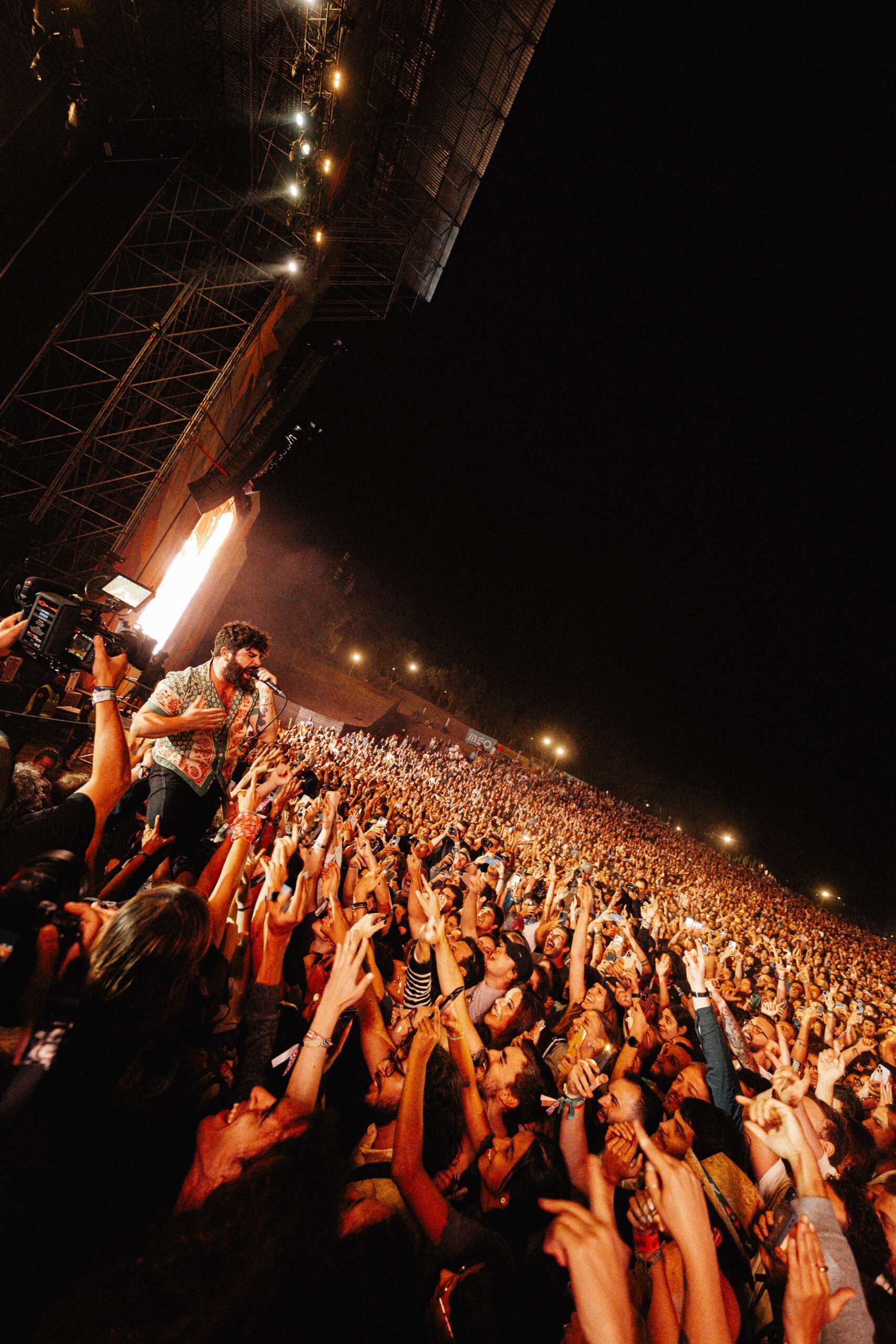 Performance ao vivo com um artista no palco, iluminação de palco, e uma multidão de fãs em um grande festival à noite.