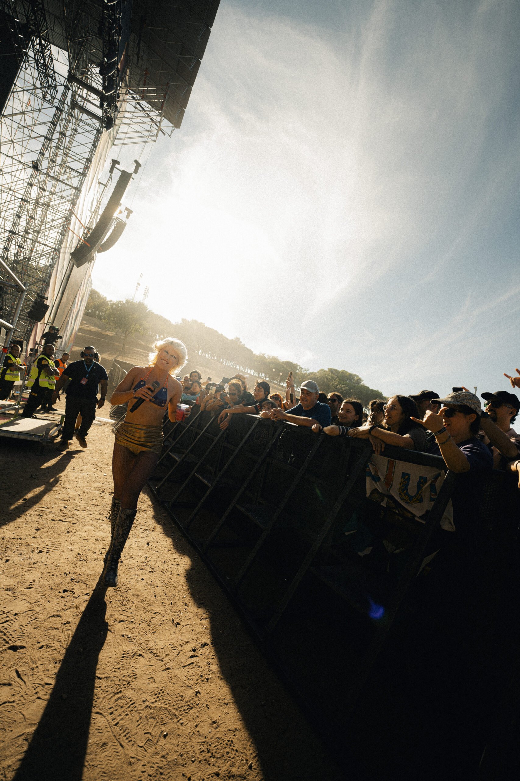 Pessoas no público assistindo a uma apresentação ao ar livre sob luz solar forte, com uma mulher caminhando na frente, segurando um copo e um microfone, e uma estrutura de palco do lado esquerdo.