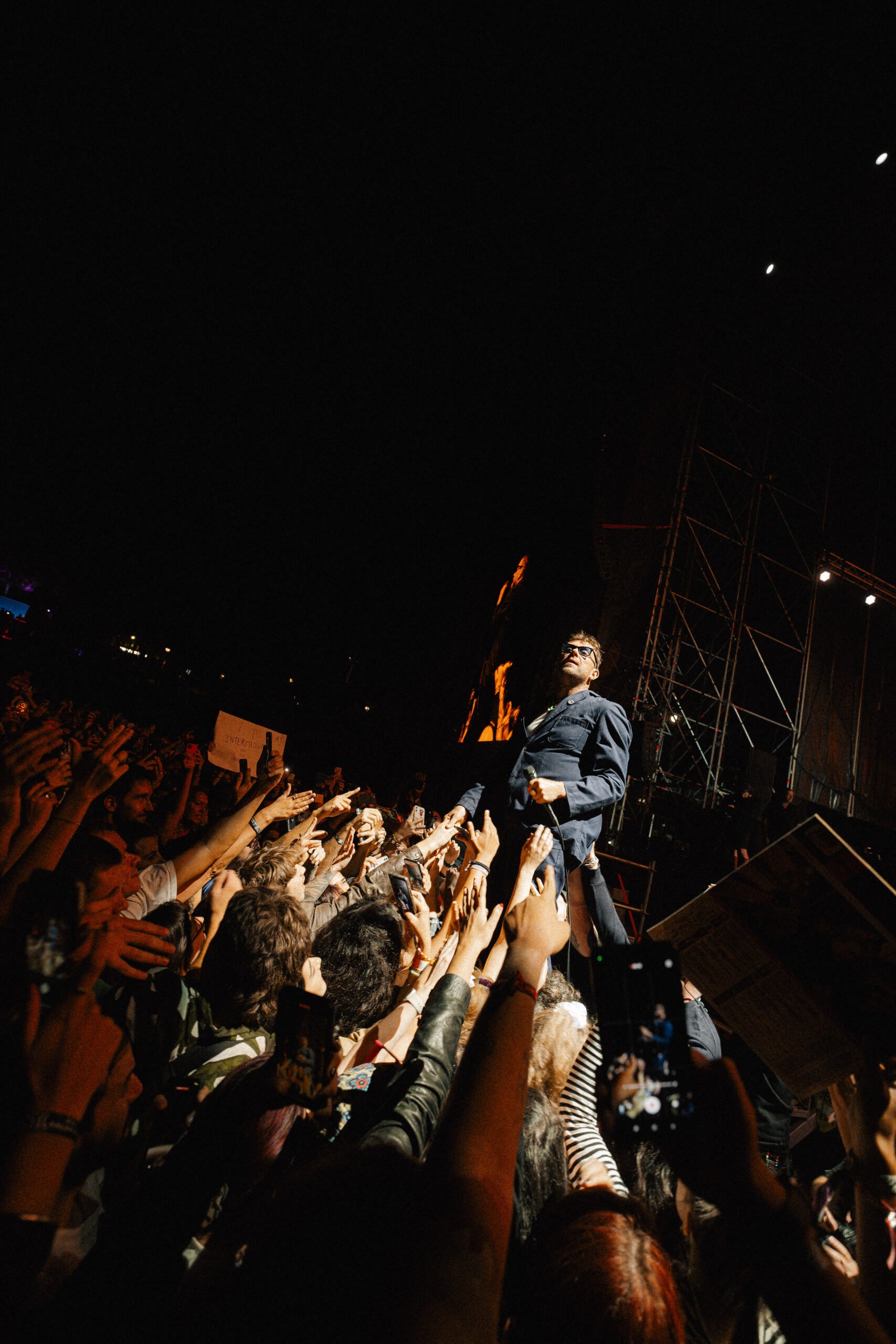 Homem de cabelo ruivo, óculos escuros e terno cinza, se apresenta em um palco durante show à noite, cercado por público que faz sinal de paz e segura celulares