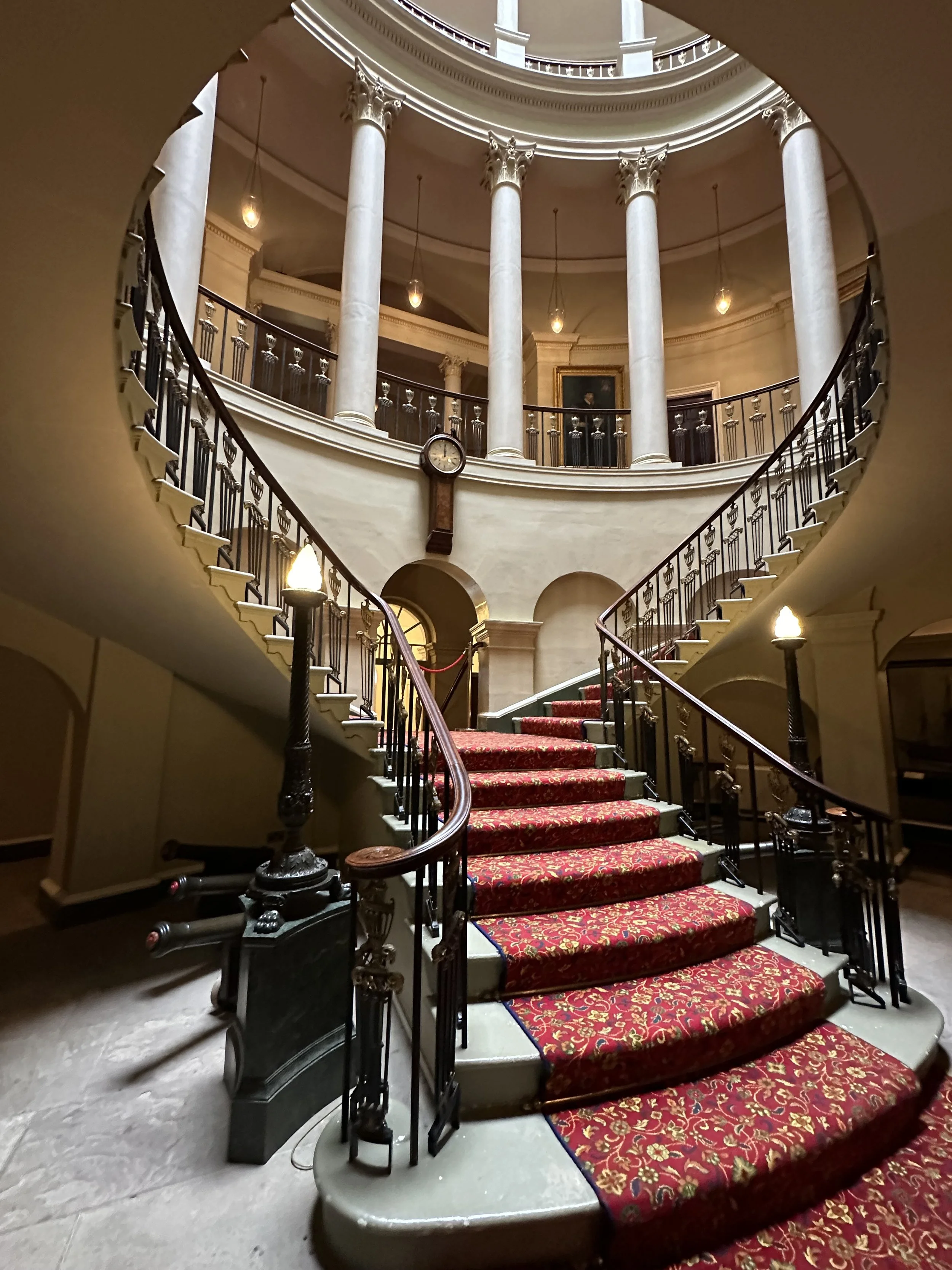 Ornate spiral staircase with red carpet, iron railings, and large columns in a historic building.