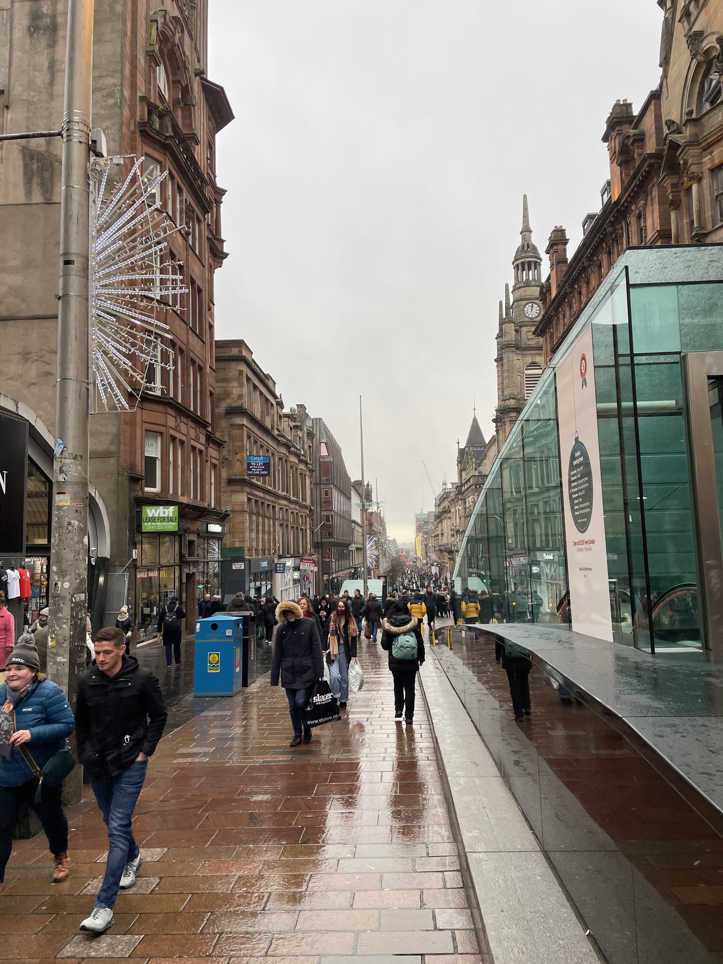 Busy urban street with pedestrians, historic buildings, and glass structure on a rainy day.