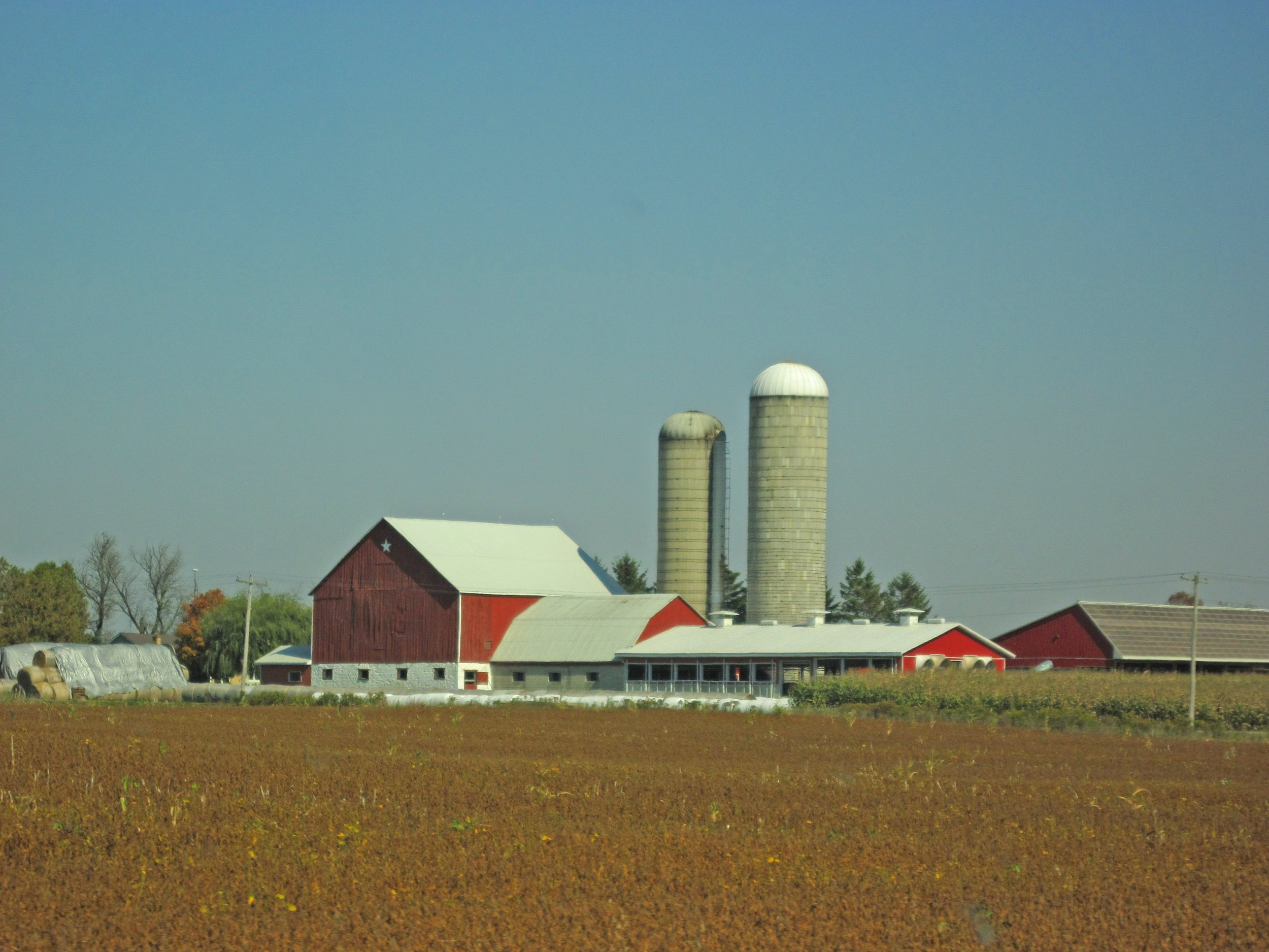 Rural farm scene with red barn, silos, and fields under a clear blue sky.
