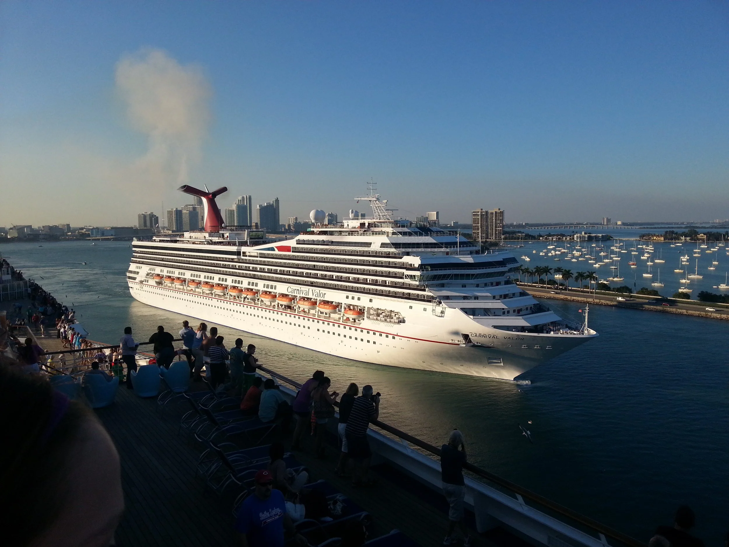 Cruise ship Carnival Valor sailing in a harbor, with people on a nearby deck watching, and a city skyline in the background.