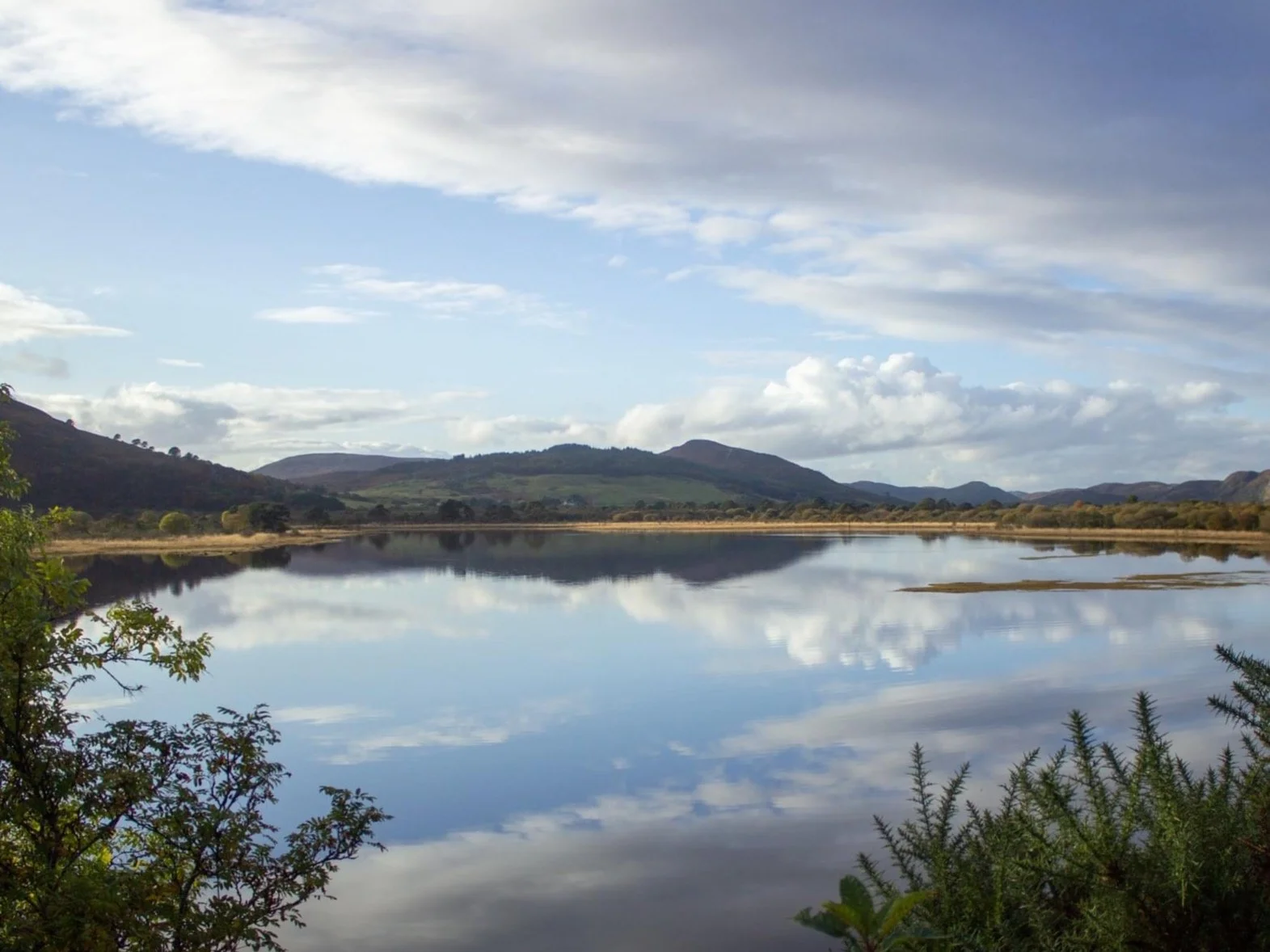 Scenic view of a calm lake reflecting blue sky and clouds, surrounded by green hills and vegetation.