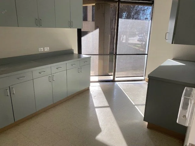 Empty kitchen with light blue cabinets, a window with a screen, and sunlight casting shadows on the floor.