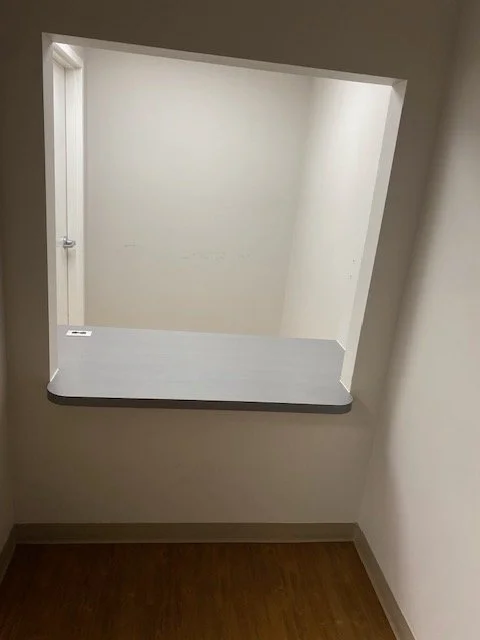 A small indoor window opening in a wall with a gray countertop below it, showing an adjacent room with light-colored walls.