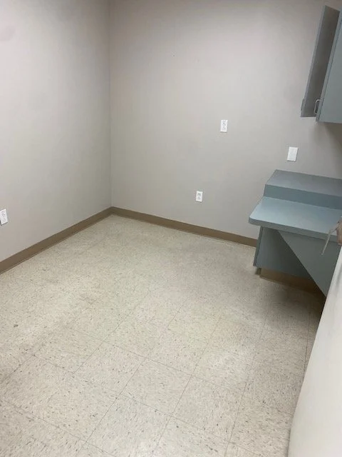 Empty room with beige walls, beige speckled floor, gray baseboards, and gray cabinets mounted on the wall.
