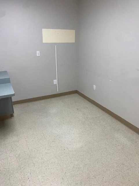 Empty room with gray walls, a small desk on the left, and a rectangular whiteboard on the back wall above a white electrical outlet, with beige baseboards and tiled floor.