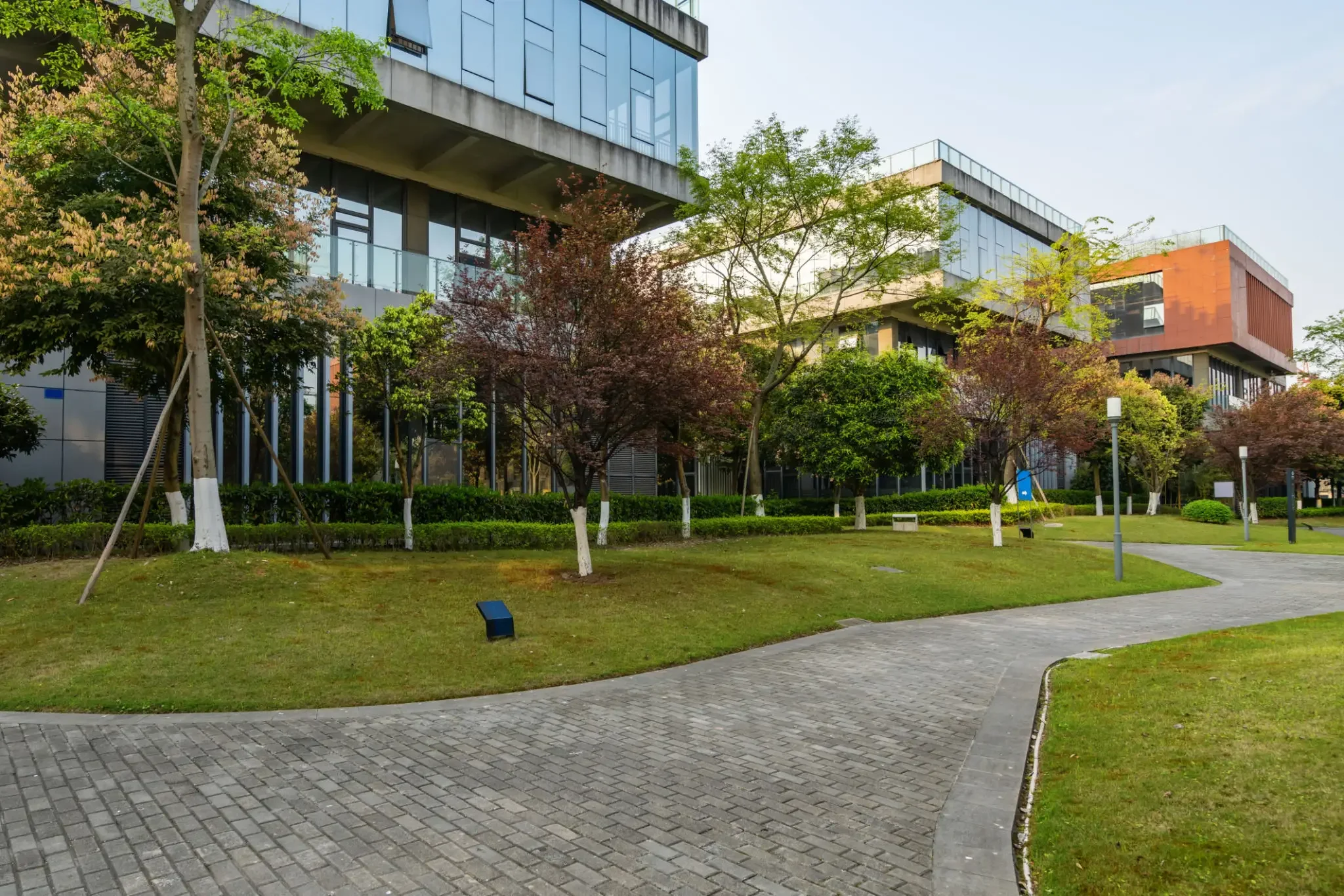 Modern office buildings with large glass windows and balconies, surrounded by a landscaped garden with trees, grass, and a winding pathway under a clear sky.