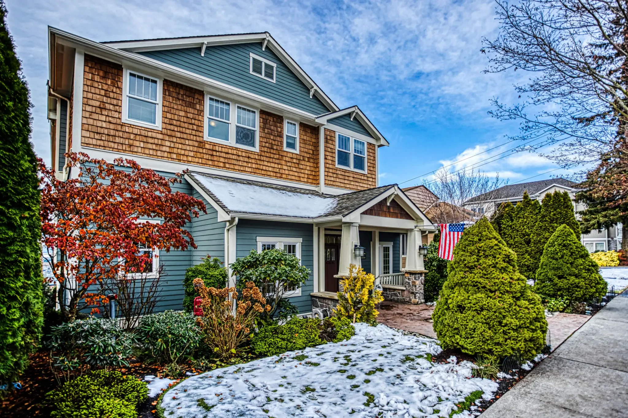A two-story house with a blue exterior, wooden shingle siding, and a stone porch, surrounded by landscaped bushes and trees with some snow on the ground and an American flag to the right.