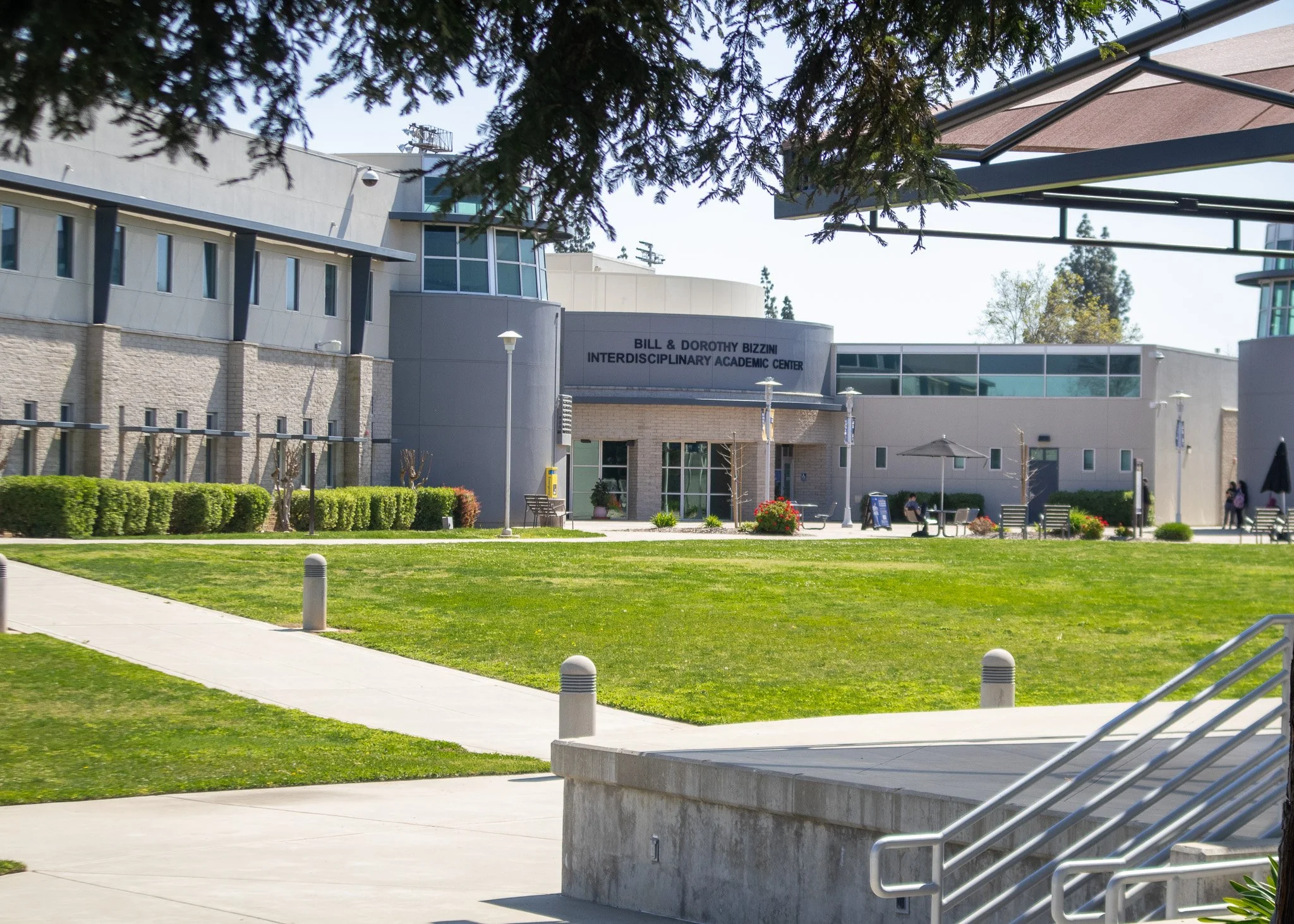 Exterior view of Bill & Dorothy Bizzini Interdisciplinary Academic Center with green lawn, pathway, benches, trees, and people walking.