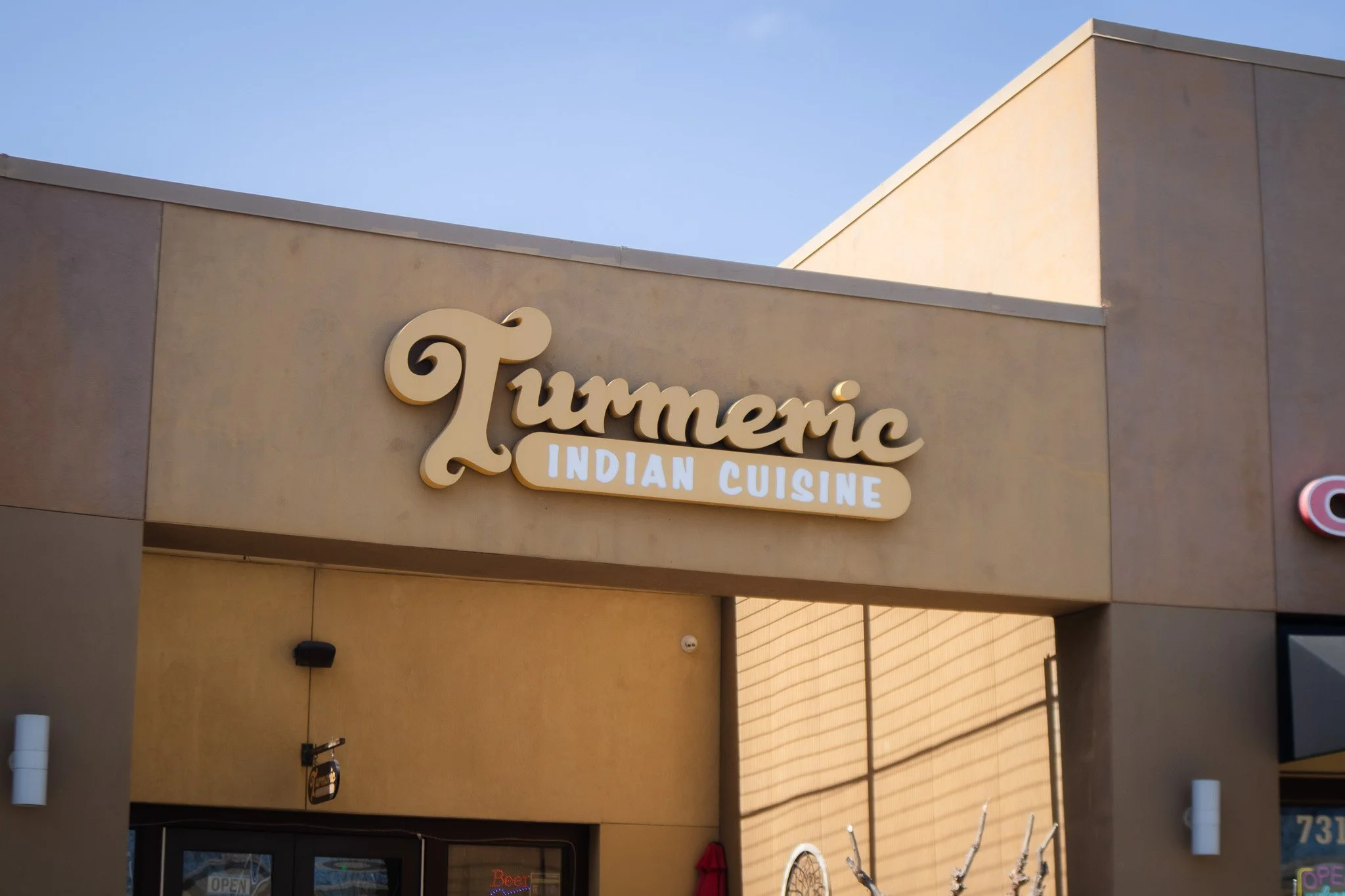 Sign on building reads 'Tummenic Indian Cuisine' with beige building exterior and clear blue sky.