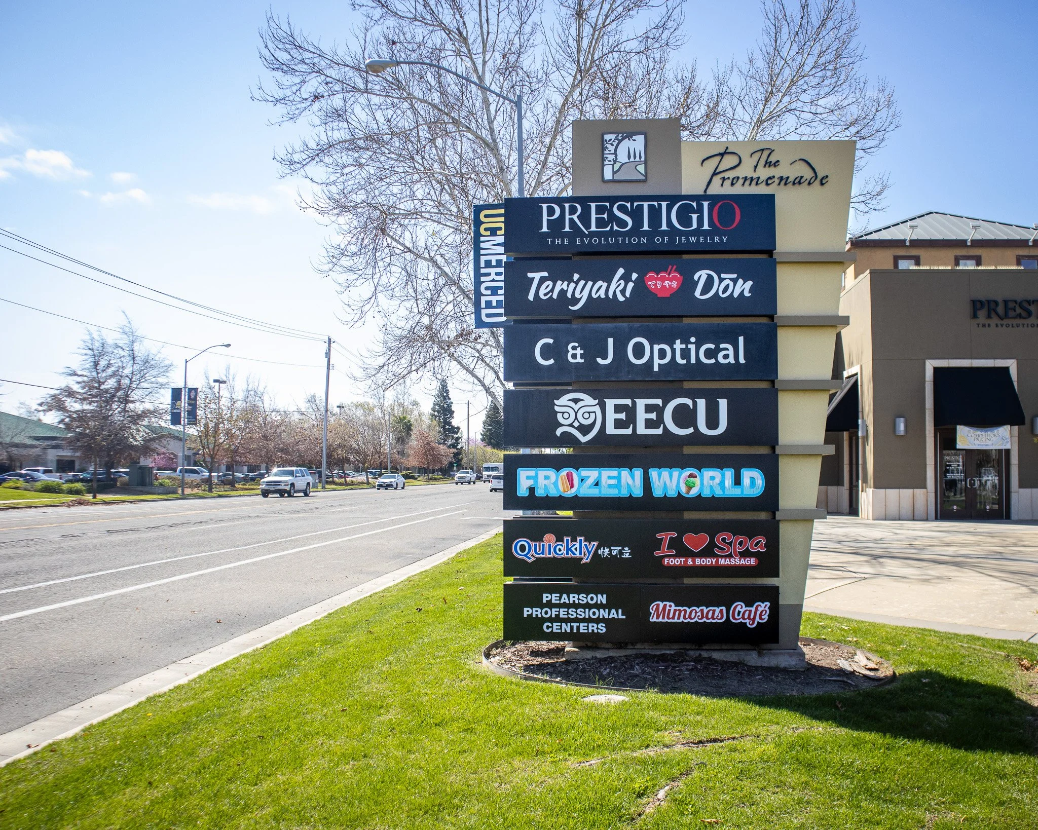 A large outdoor signs with multiple business names on a sidewalk grassy area, including Prestige, Teriyaki Don, C & J Optical, ECU, Frozen World, Quickly, Spa, Pearson Professional Centers, and Minosas Café.