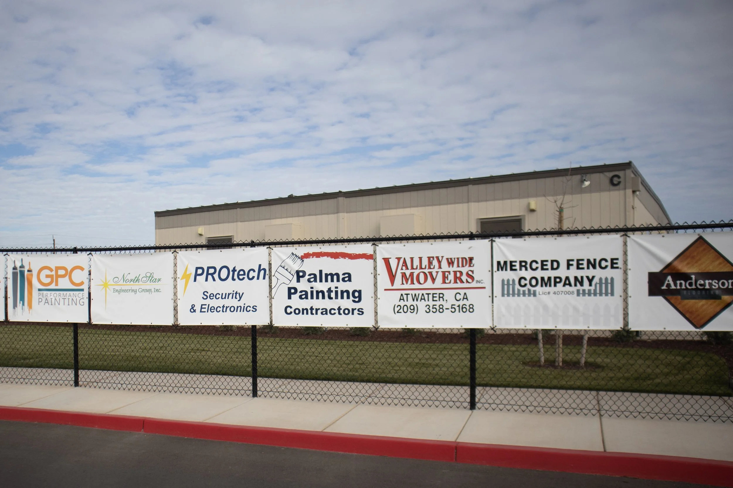 Multiple business banners hung on a chain-link fence in front of a beige industrial building, advertising services like painting, security electronics, moving, fencing, and construction company.