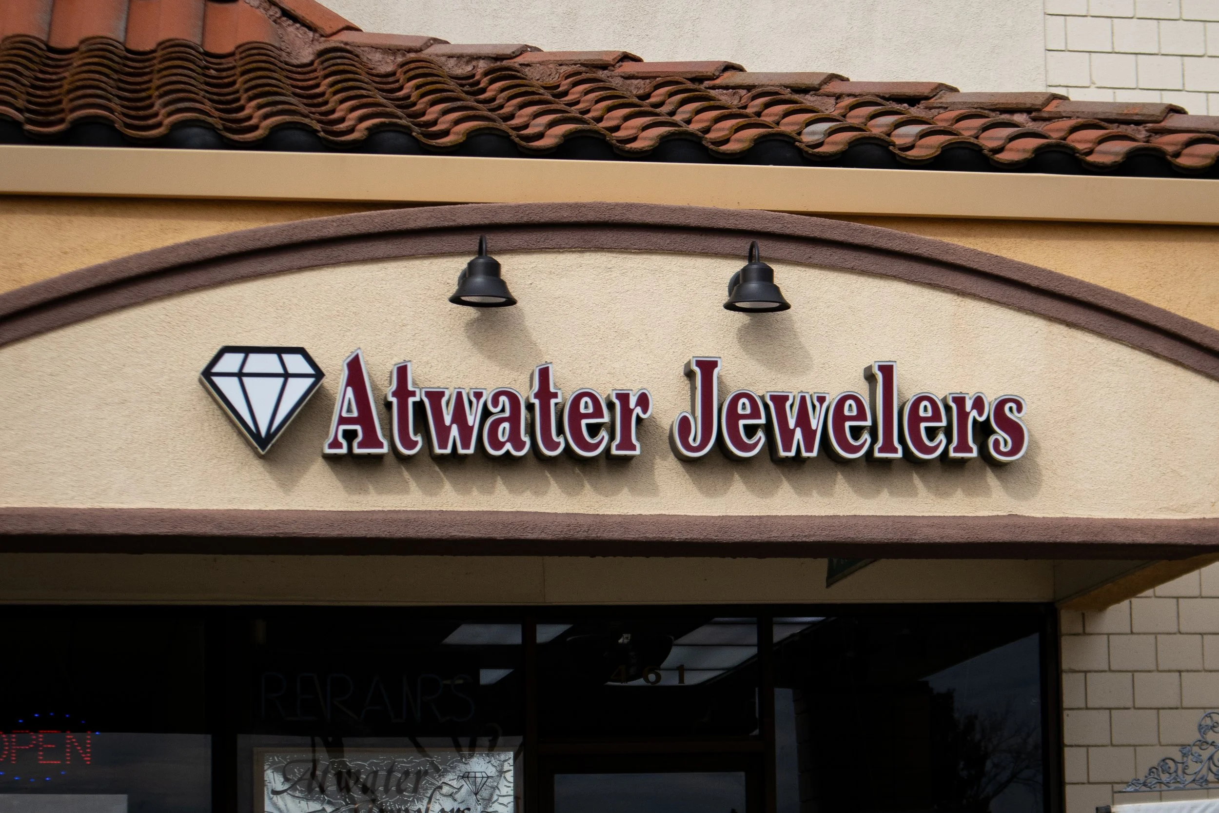 Storefront sign for Atwater Jewelers with a diamond icon on a beige wall, illuminated by two black outdoor lights.