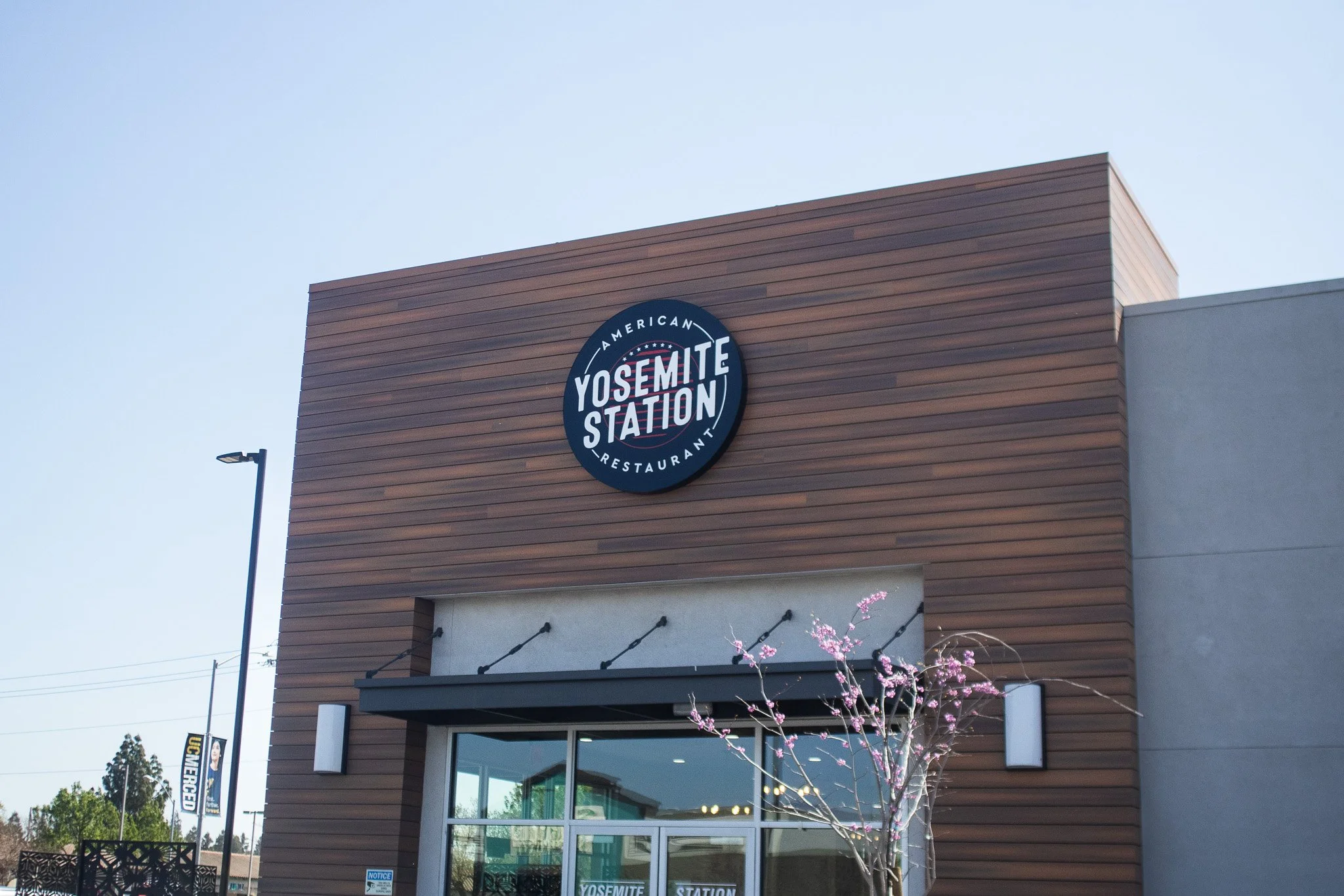 The exterior of a restaurant named Yosemite Station with a wooden facade, glass entrance, and a lit sign above the door. A small tree with pink blossoms is in front.