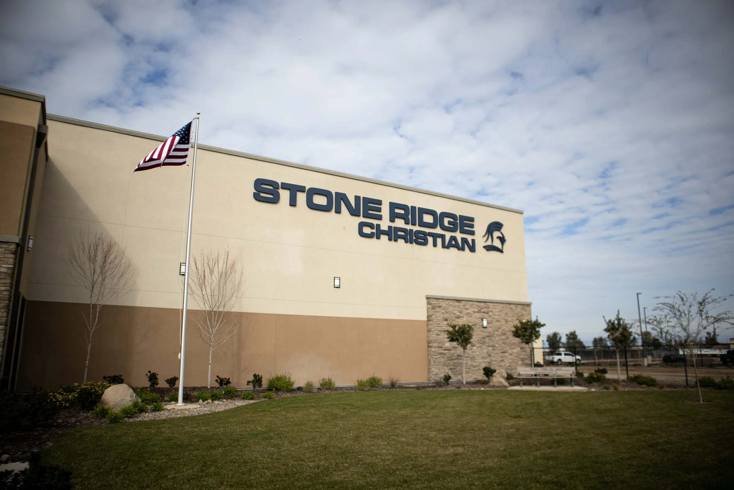 Exterior of Stone Ridge Christian school with American flag, shrubs, trees, and a bench in the foreground under a cloudy sky.