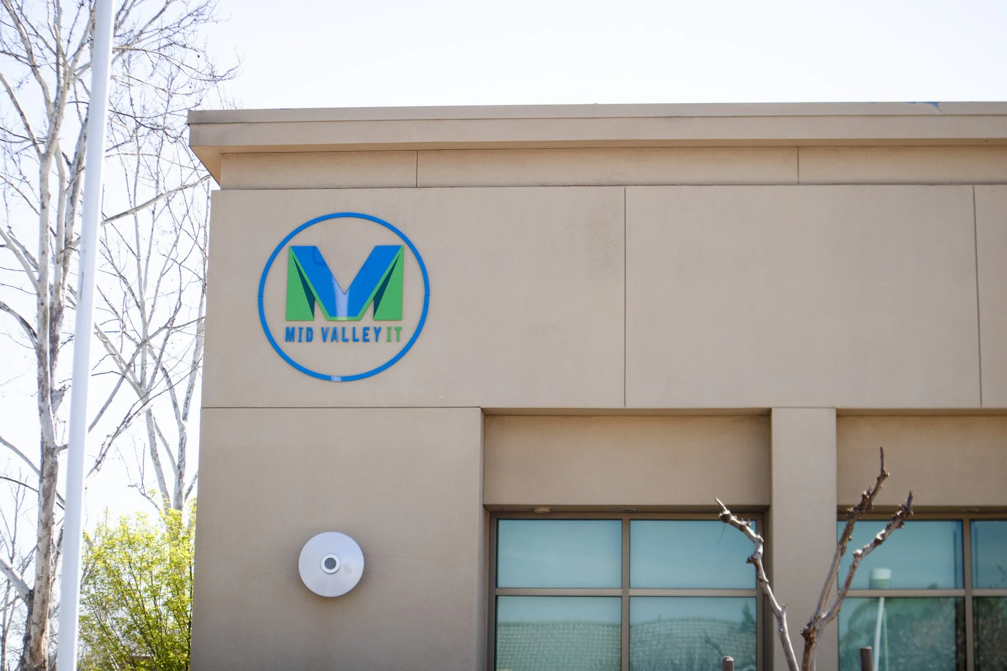 Exterior of a building with a sign that reads 'MID VALLEY IT' and a large circular logo featuring the letter M in blue and green.