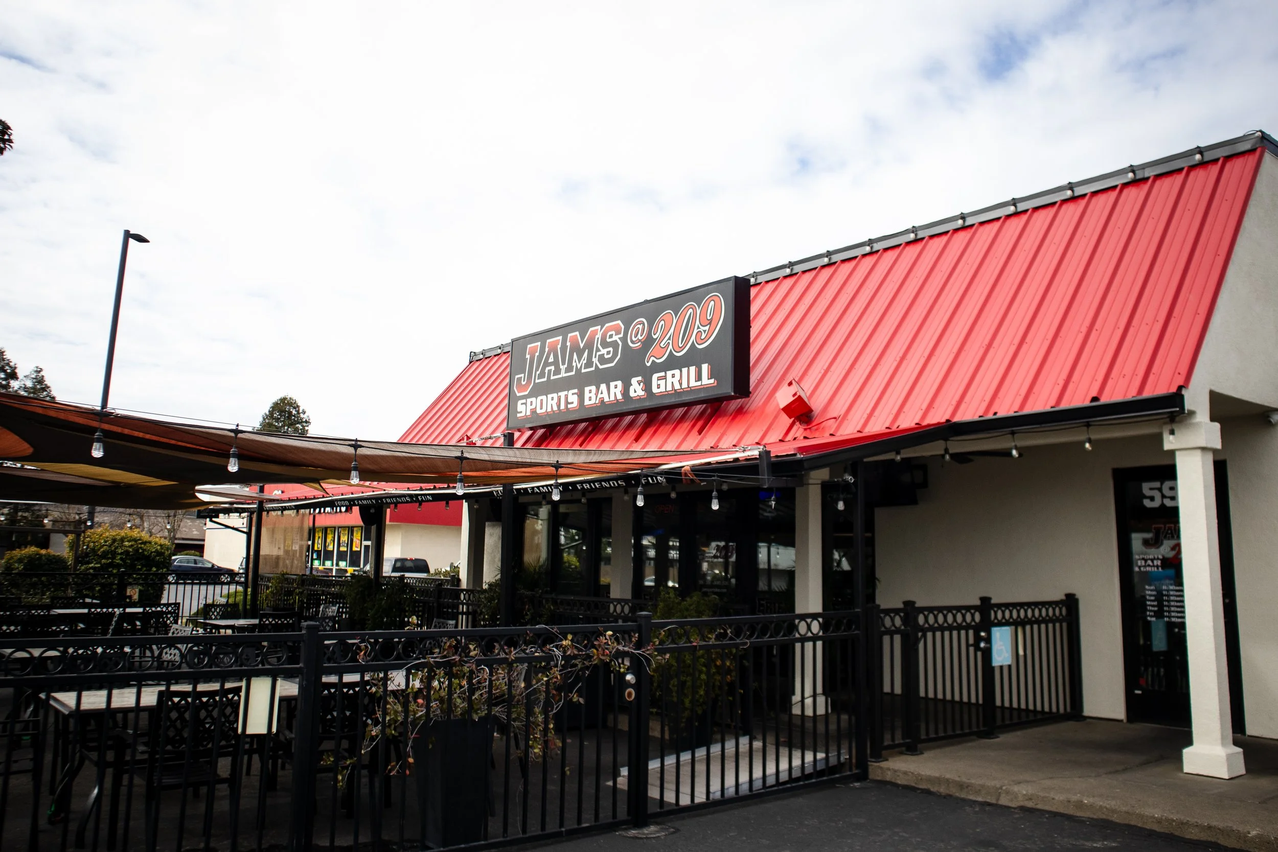 Exterior view of JAMS Sports Bar & Grill with a red sloped metal roof and a black sign displaying its name, featuring outdoor seating area with tables, chairs, and string lights.