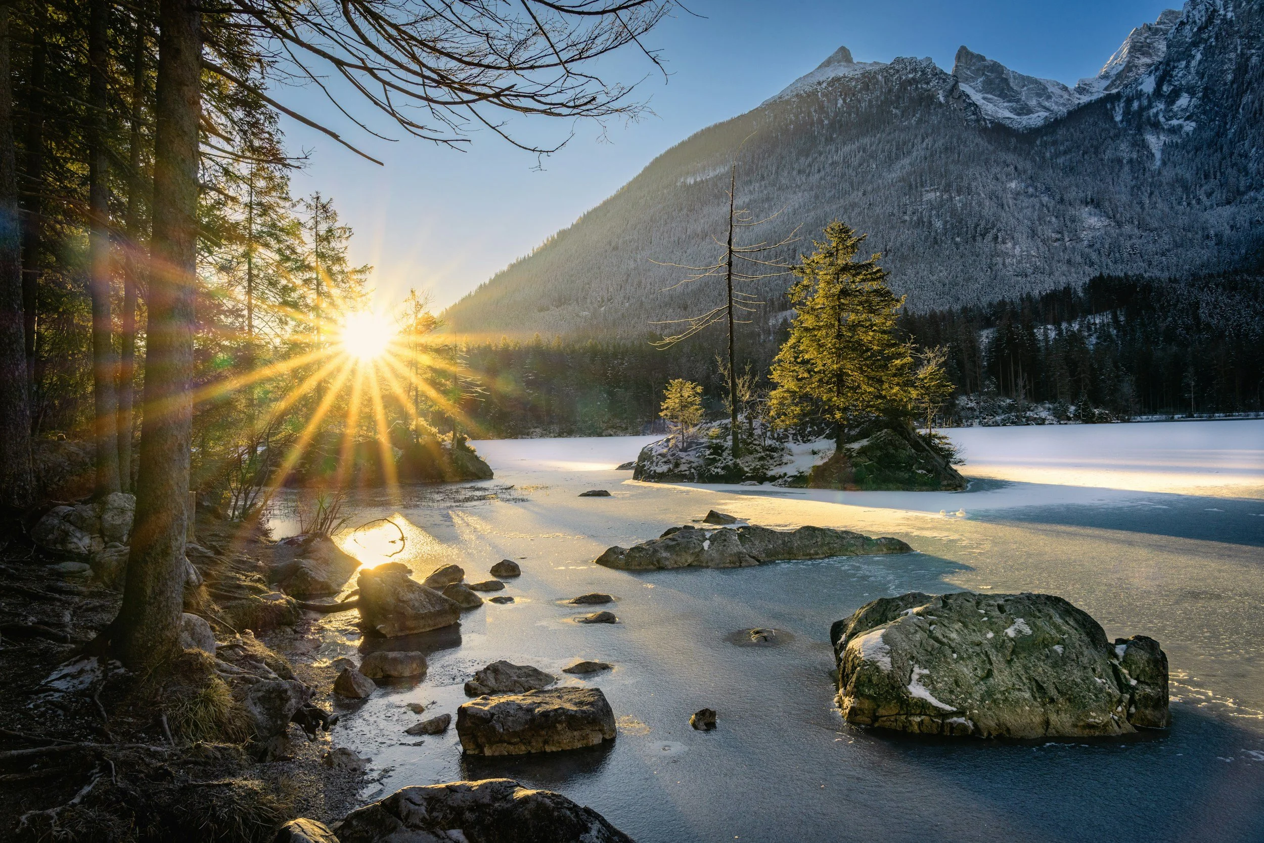 Sunrise over a snow-covered lake with trees and mountains in the background.