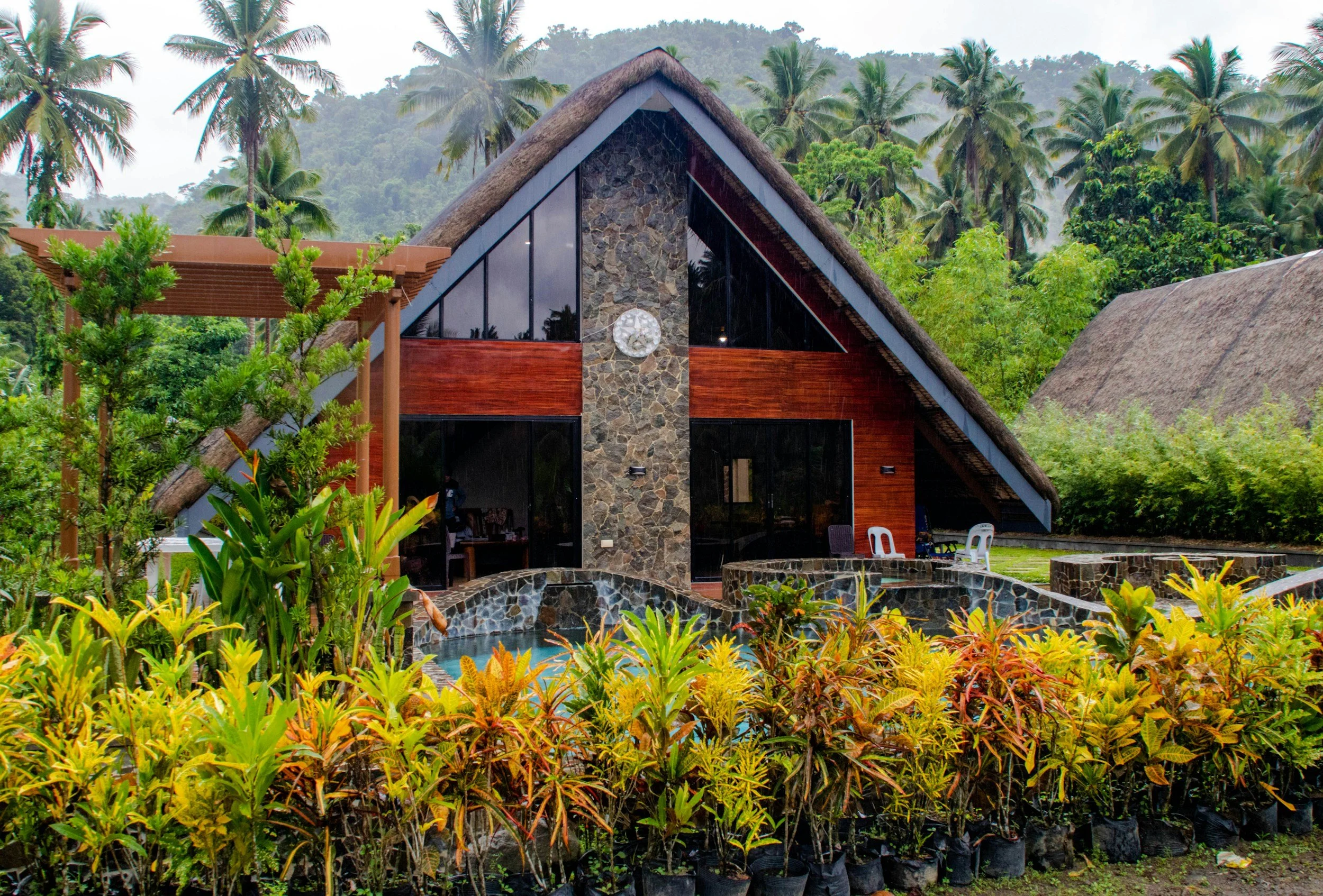 A modern A-frame house with a stone and wooden exterior, large glass windows, a surrounding lush tropical garden, and a hill with palm trees in the background.