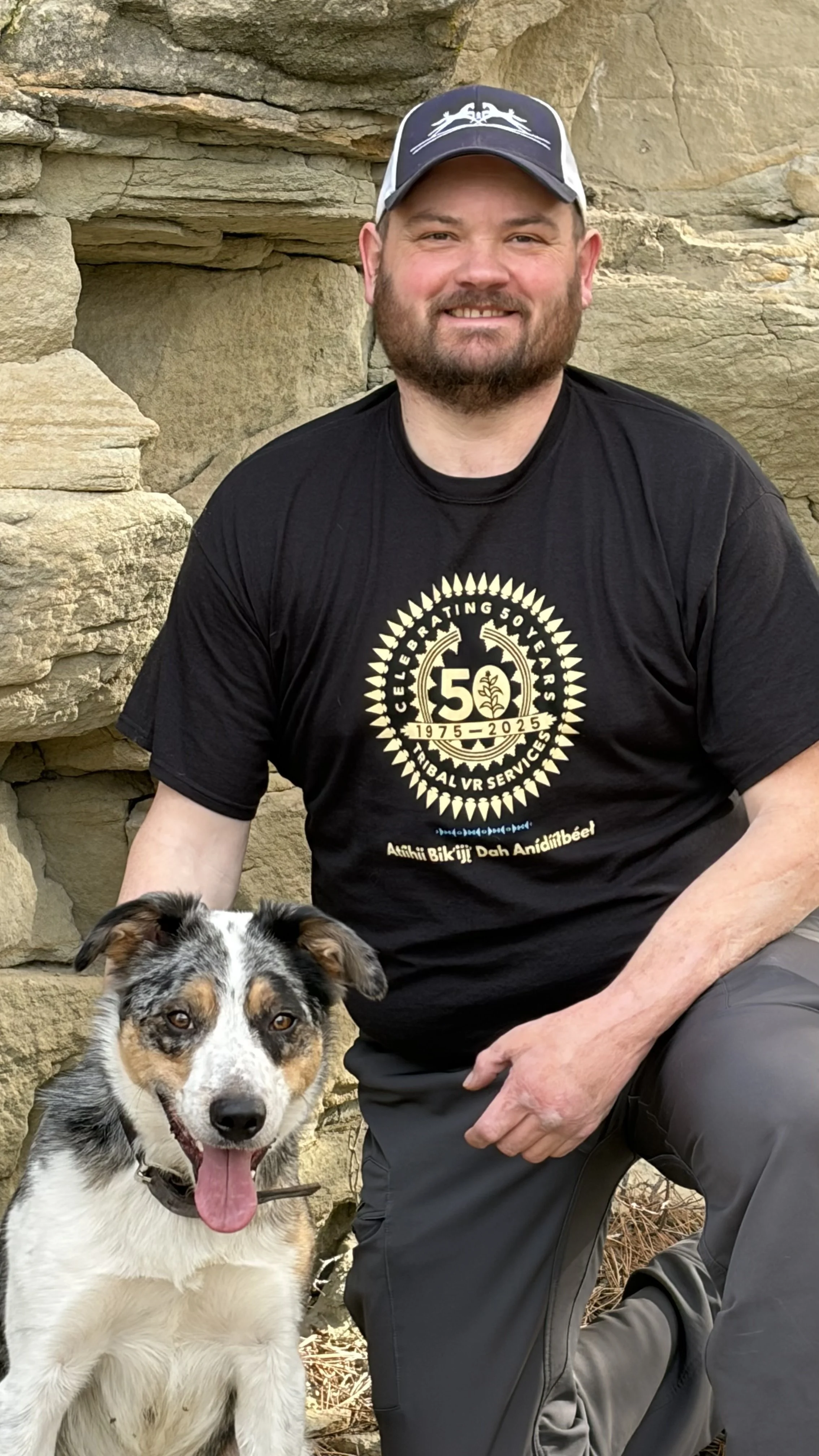 A smiling man with a beard wearing a camouflage hat and outdoor gear, outdoors in a natural setting.