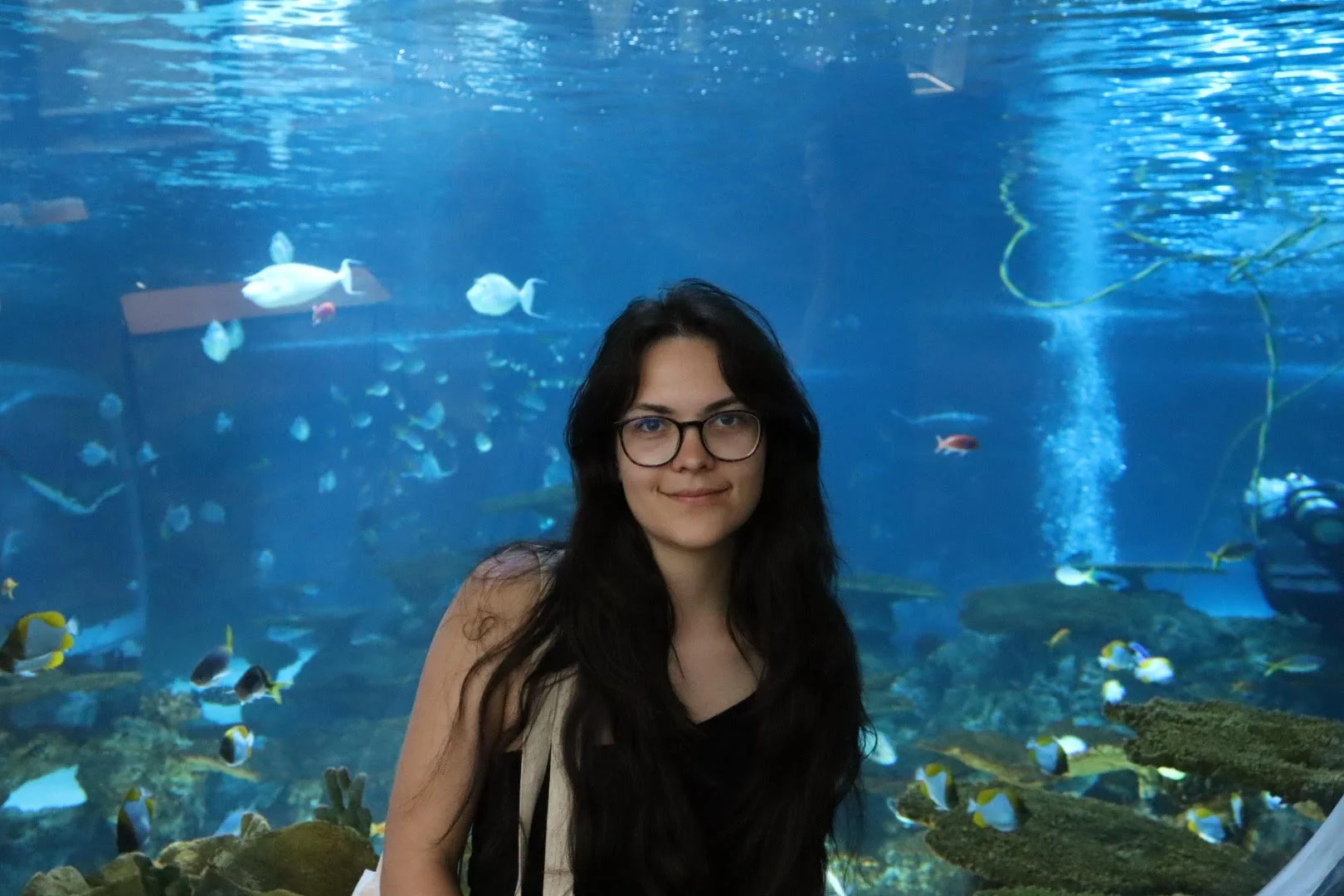A woman with long dark hair and glasses standing in front of an aquarium tank filled with various colorful fish and aquatic plants.