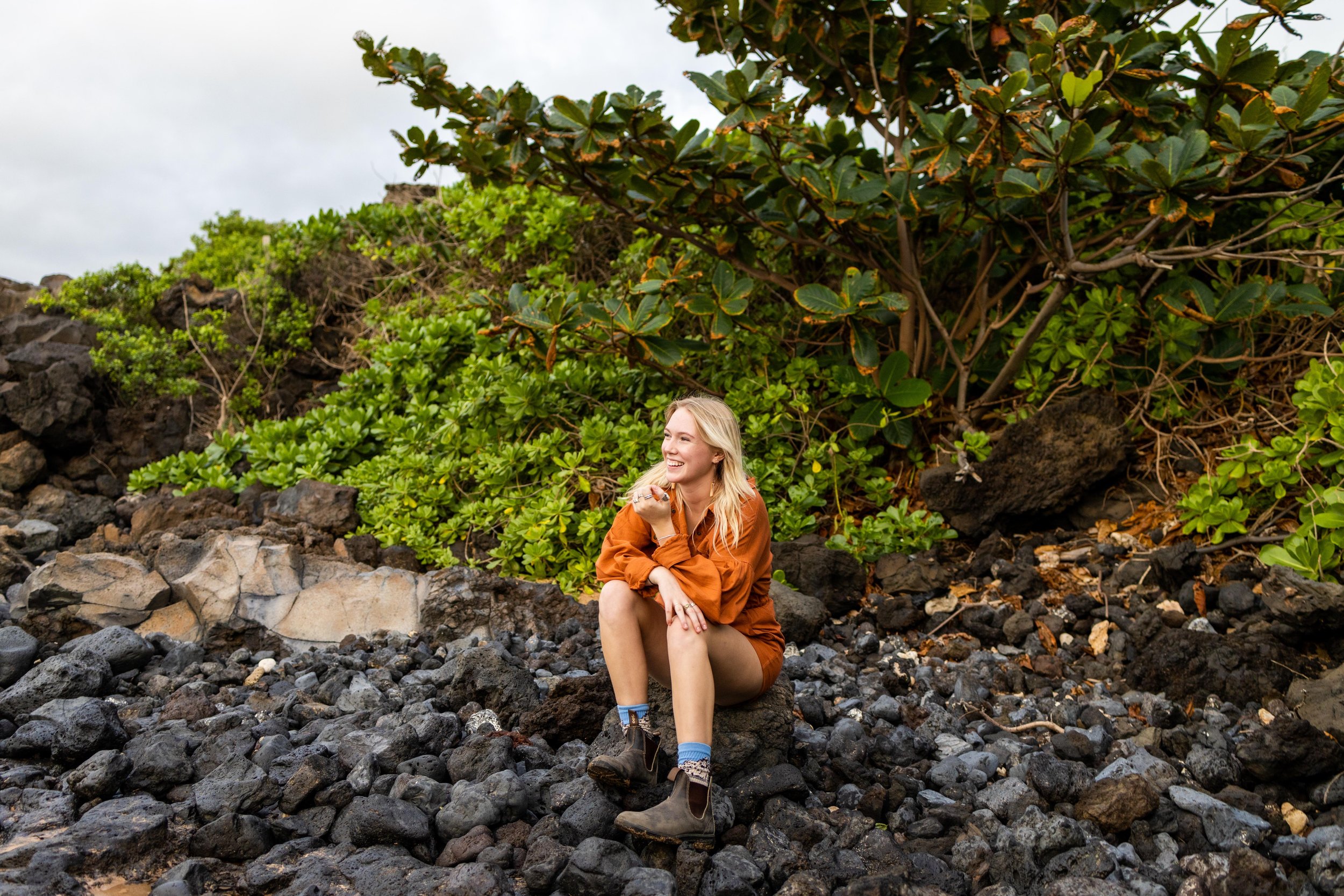 A woman in an orange jacket and shorts sitting on black volcanic rocks near green shrubbery and trees.