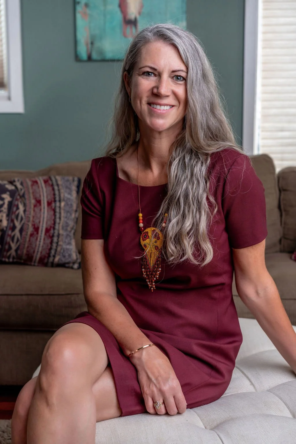 A woman with long gray hair sitting on a white ottoman in a living room, smiling, wearing a maroon dress and a colorful beaded necklace.