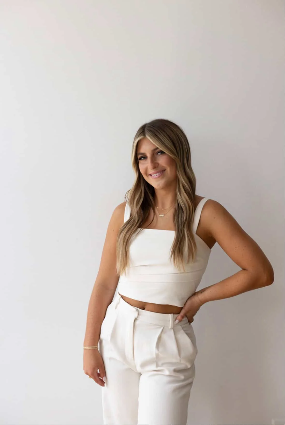 A woman with long, wavy blonde hair posing in front of a plain, light-colored background. She's wearing a white crop top and high-waisted white pants, smiling softly with her hand on her hip. Hairstylist now serving Perrysburg, Toledo, Sylvania, Ohio