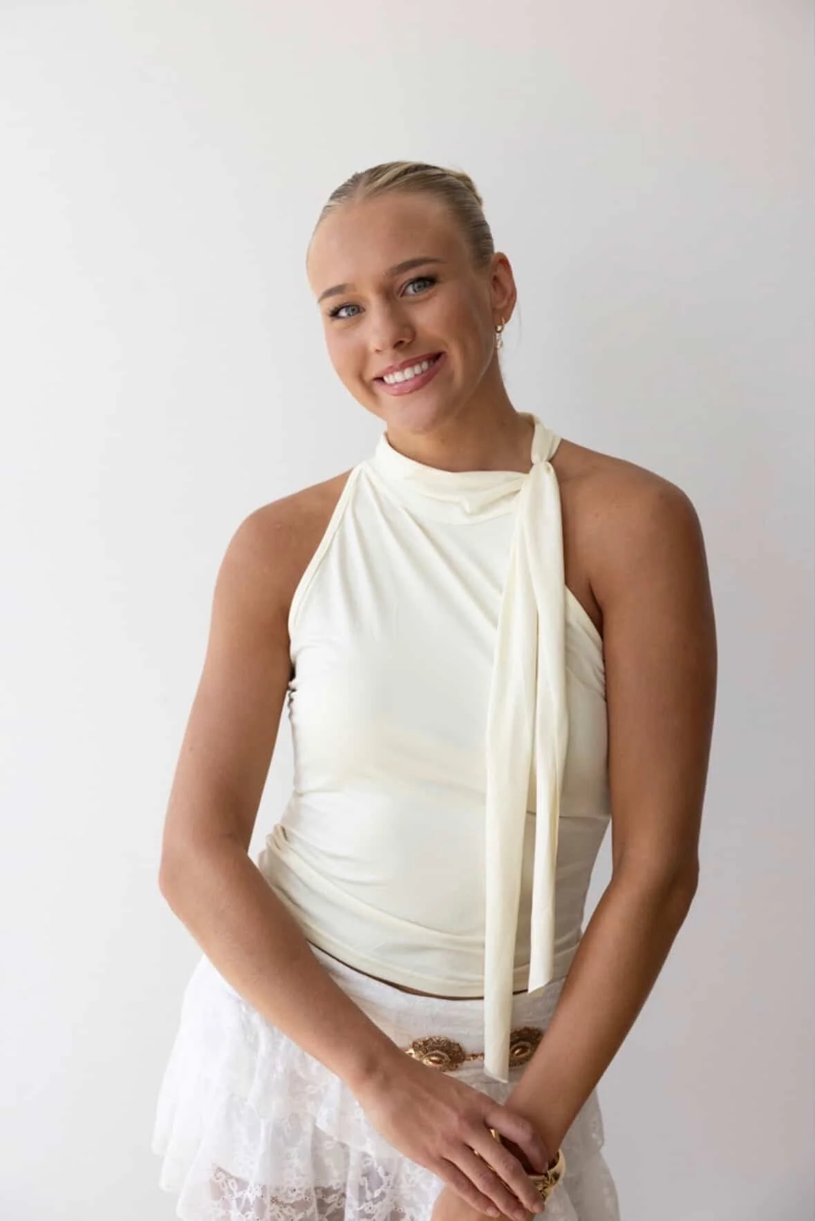A young woman with blonde hair in a sleek bun, smiling, wearing a sleeveless ivory top with a necktie detail and a white lace skirt, standing against a plain white background. Hairstylist now serving Perrysburg, Toledo, Sylvania, Ohio