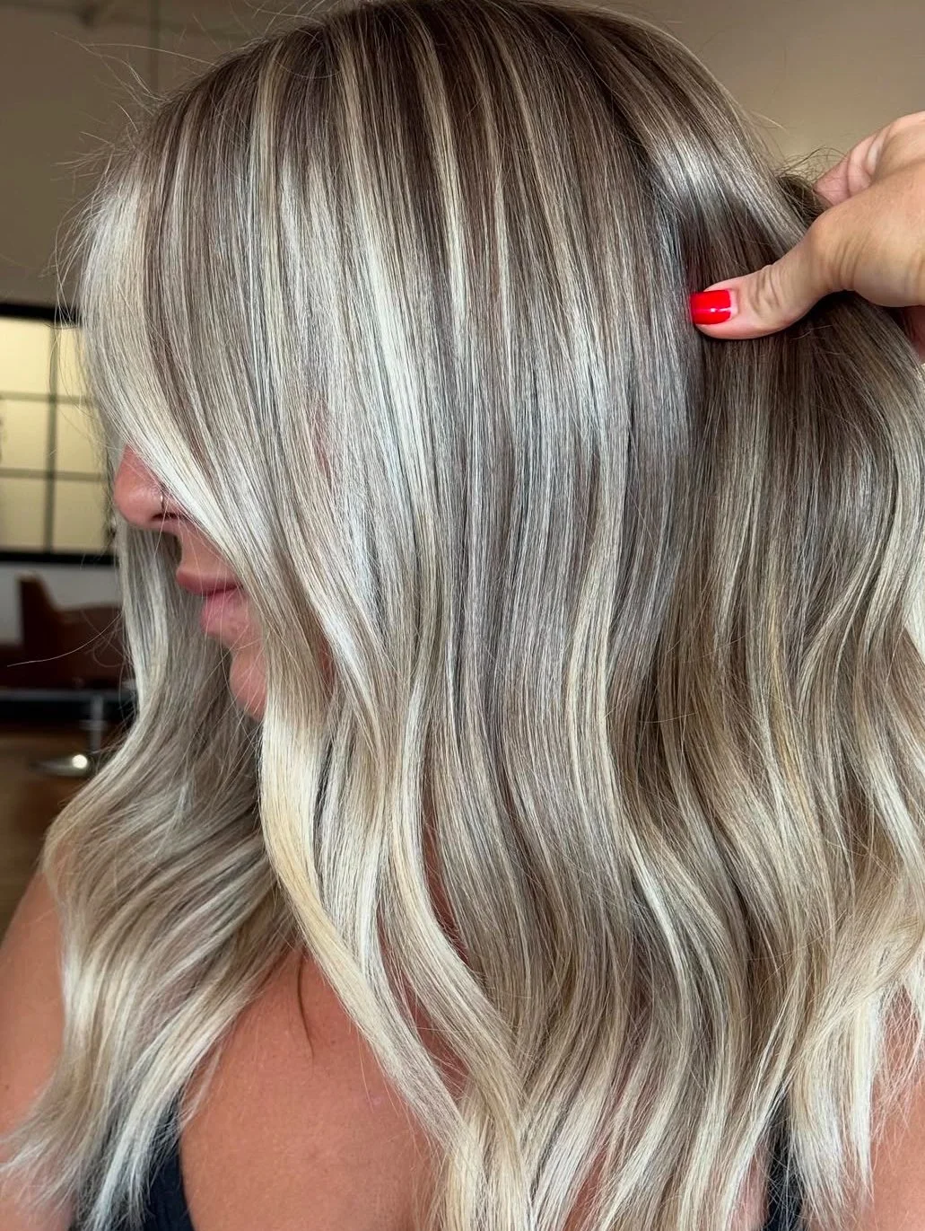 Close-up of a woman with long, wavy, blonde hair with highlights, being styled by a hand with red nail polish.