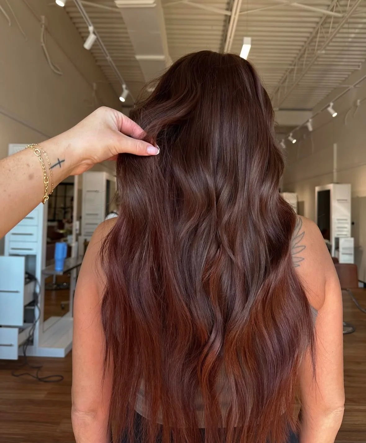 A woman with long, wavy, brown hair in a hair salon, with a stylist's hand holding a section of her hair.