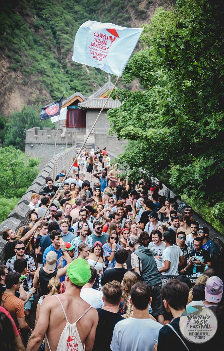 Crowd of people attending a music festival at the Great Wall of China, with flags and greenery in the background.