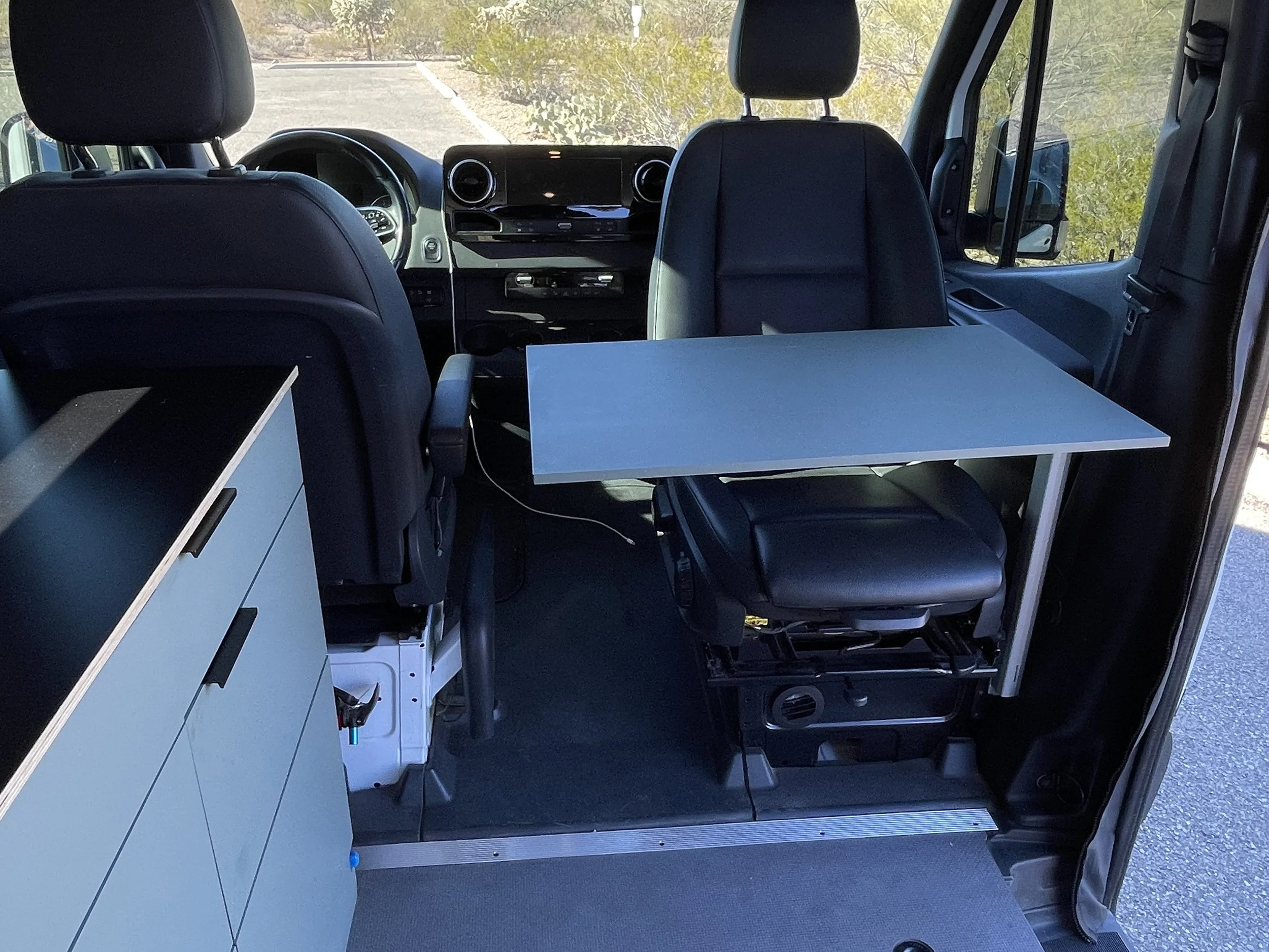Interior of a camper van showing the driver and passenger seats with a swivel table, storage cabinet on the side.