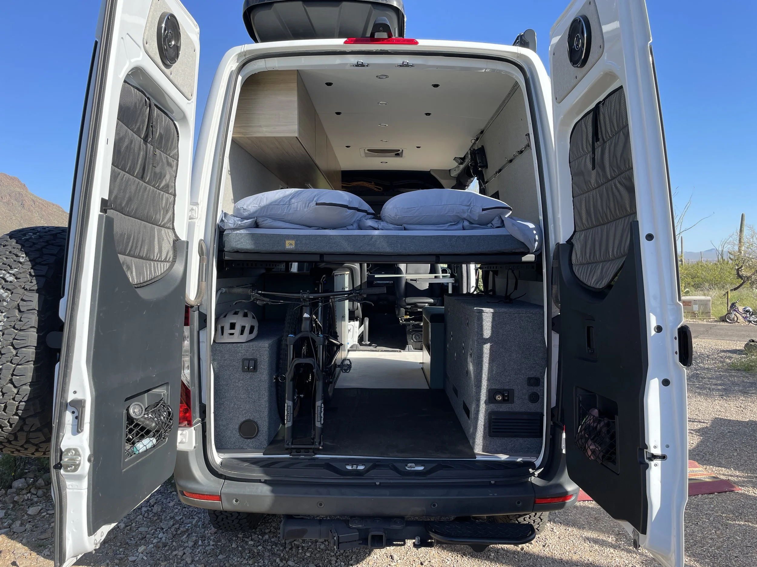 Interior view of a camper van with open rear doors, showing a bed with pillows, stored bicycles, overhead cabinets, and a spare tire attached to the door.