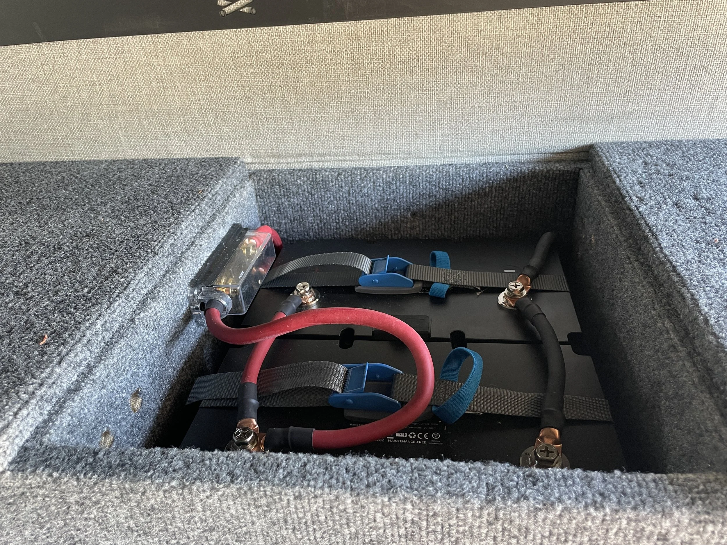 Car battery setup with red and black cables inside a carpeted enclosure, featuring a fuse holder and securing straps.